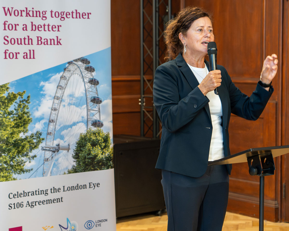 Cllr Claire Holland into a microphone at a lectern during an event, with a banner beside her reading “Working together for a better South Bank for all” and referencing the London Eye S106 agreement.