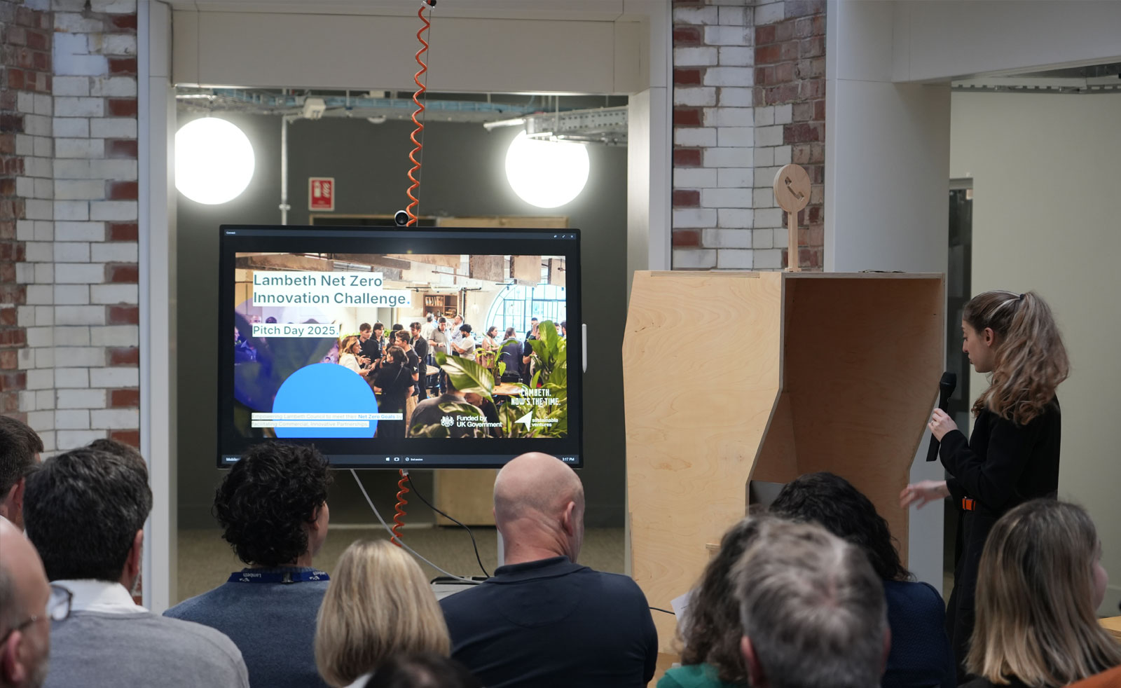 An audience sits in rows facing a screen which reads 'Lambeth Net Zero Innovation Challenge, Pitch Day 2025". A speaker stands on the right of the image with a microphone in hand facing the screen.