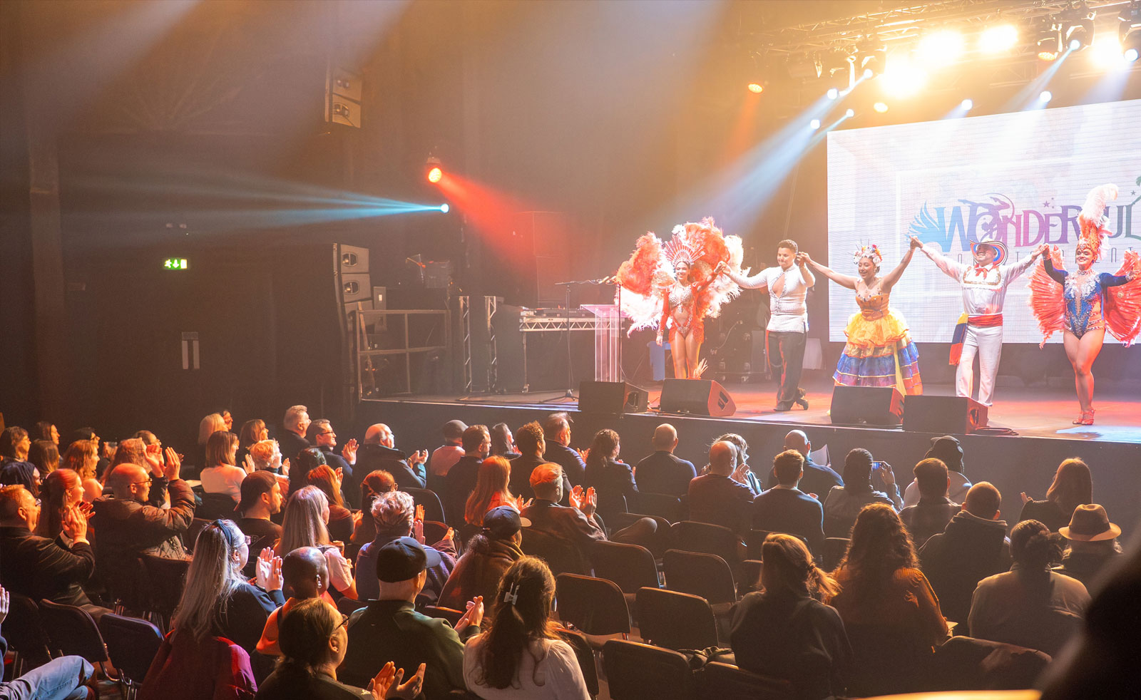 Performers dressed in traditional Latin American dance costumes, performing on a stage in front of a seated audience