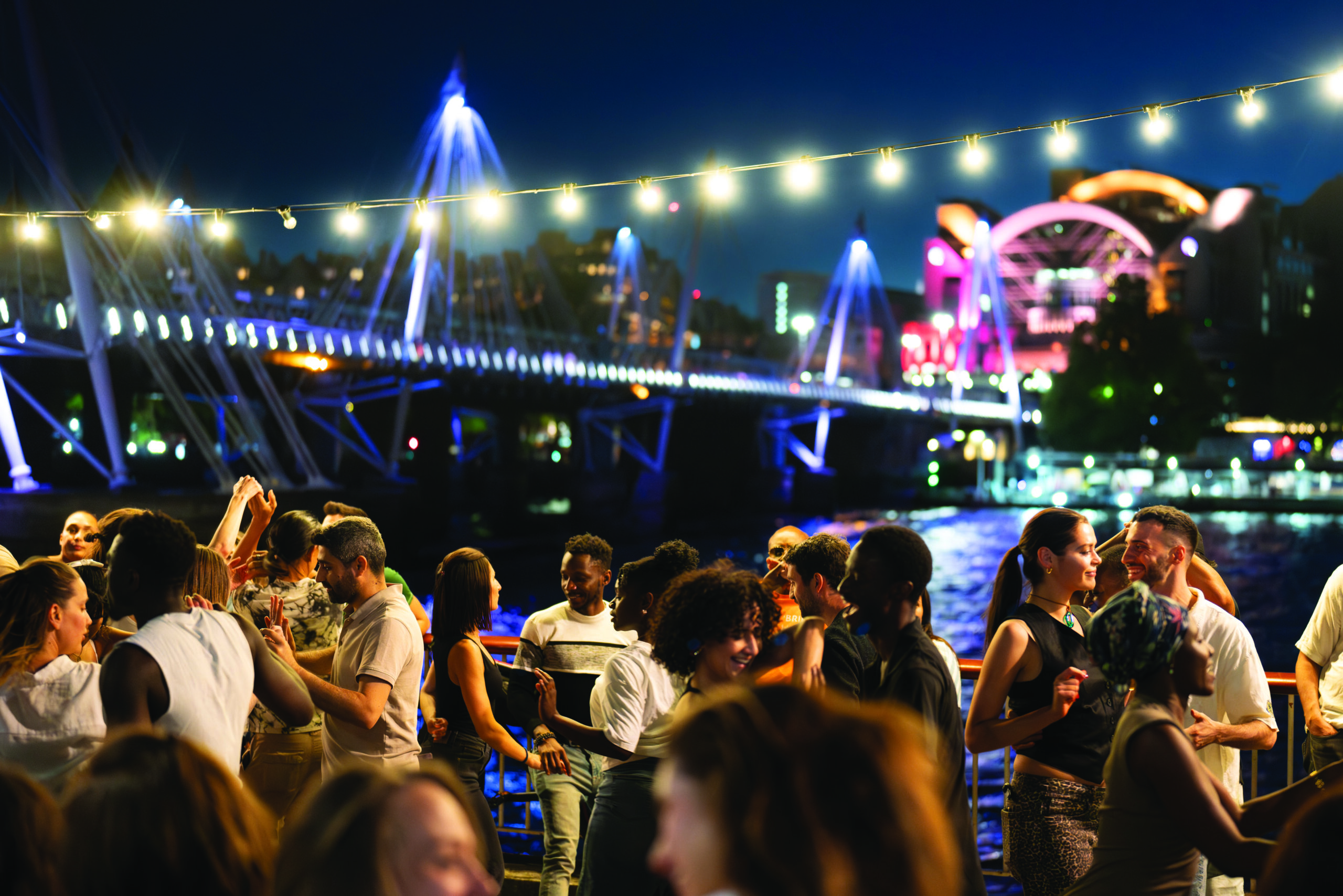 A large group of people standing and talking in an outdoor nightlife space along the river on South Bank