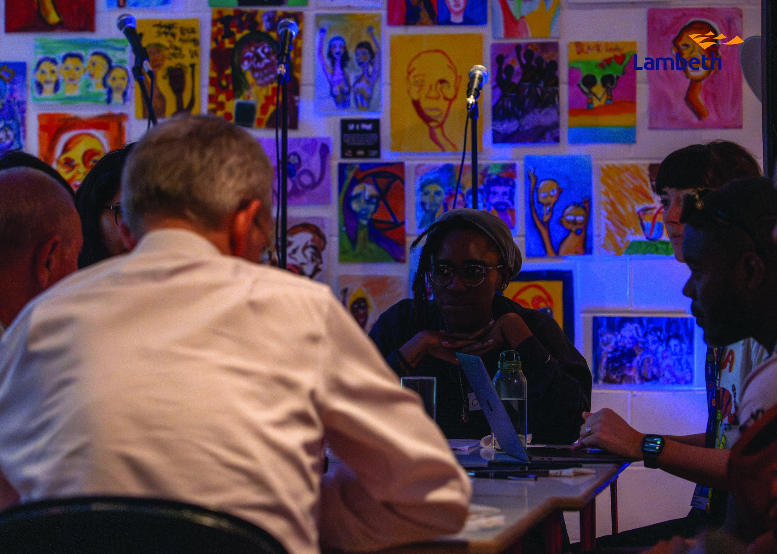 A group of people sit around a table in conversation in front of a wall of community artworks