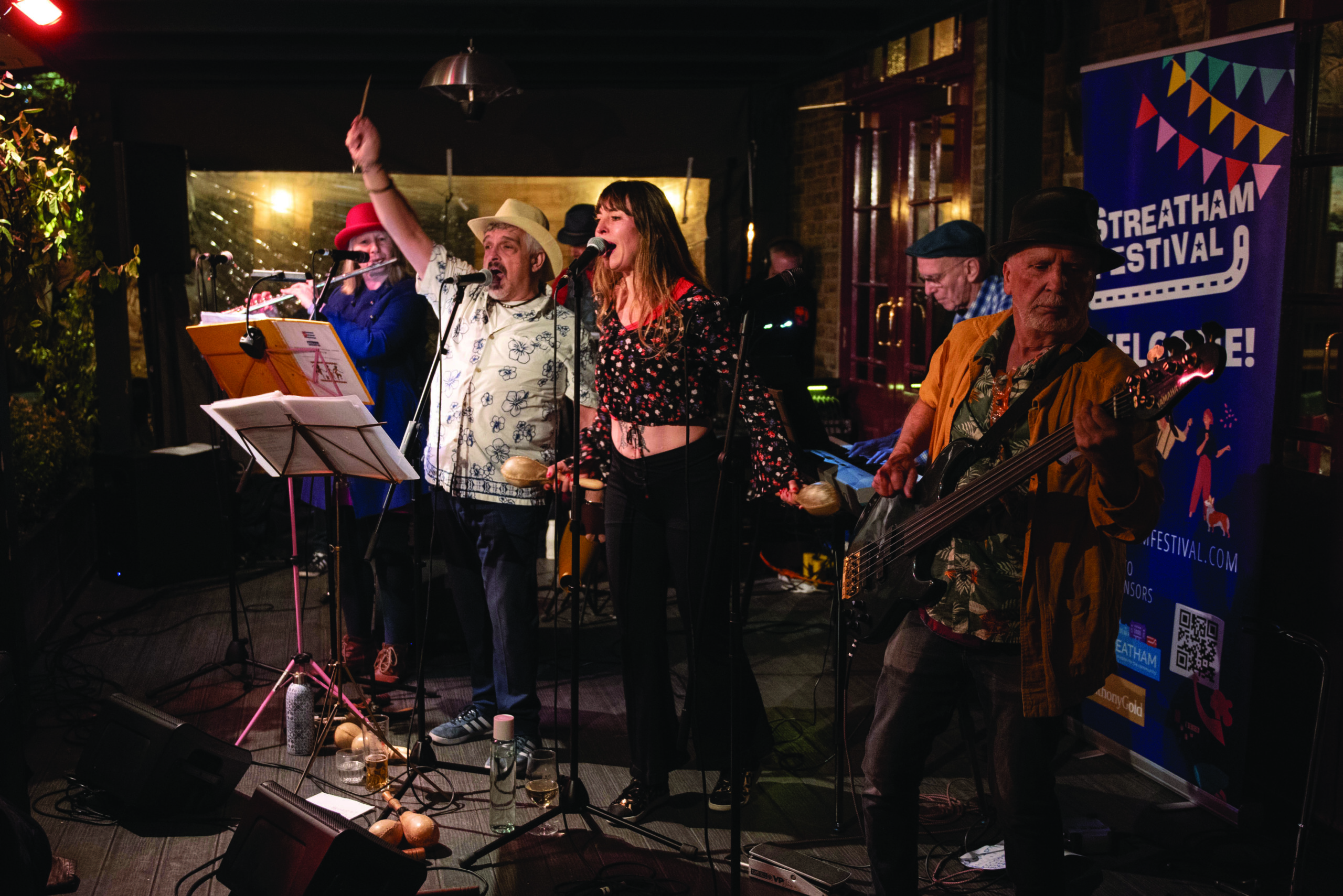 A band of people playing different instruments, led by two front people singing into microphones, performing in a bar