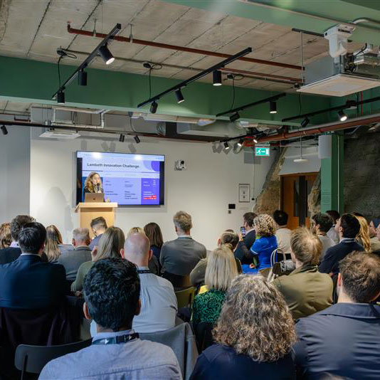 An audience seated in a conference room listens to a speaker presenting at a podium, with a presentation slide displayed on a screen at the front of the room.