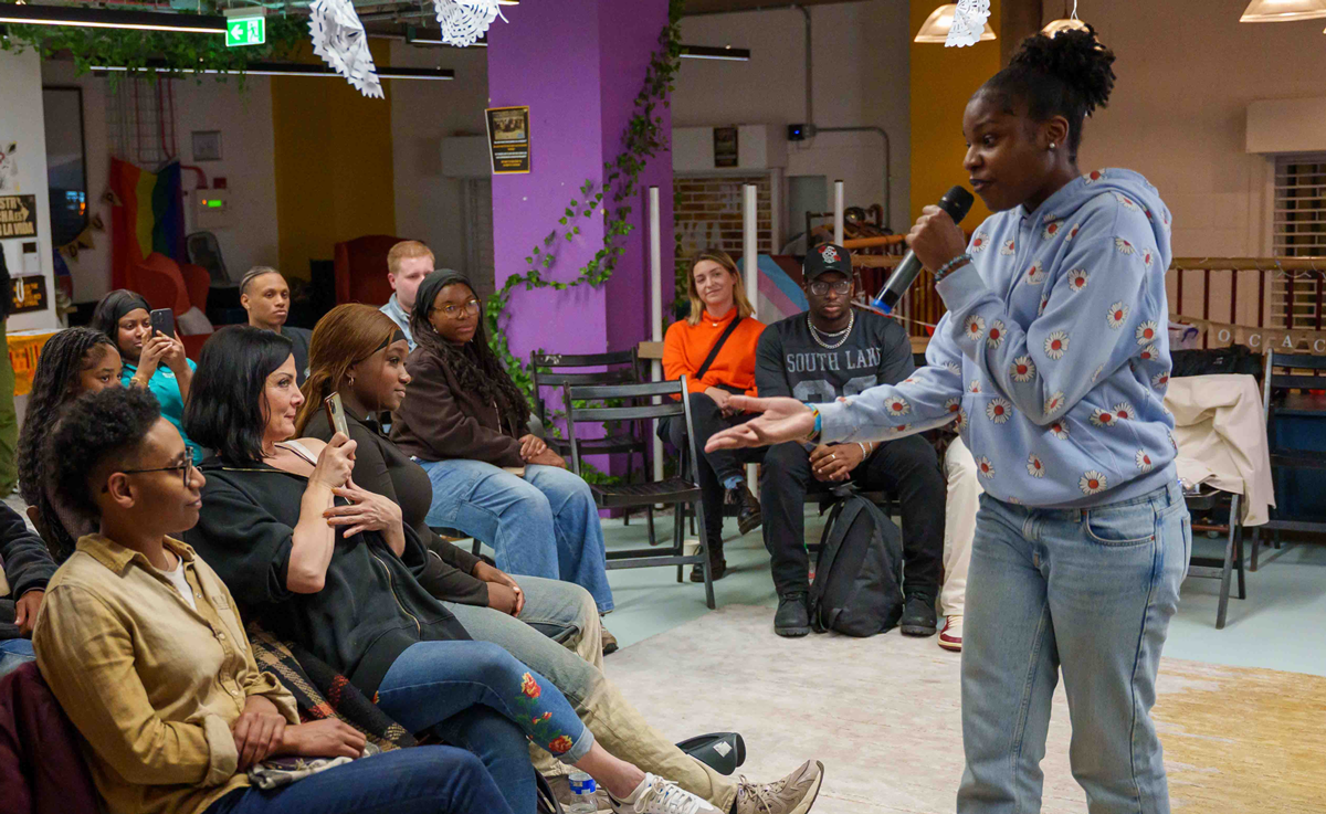 A young person in a blue sweatshirt singing into a mic and reaching out towards a seated audience