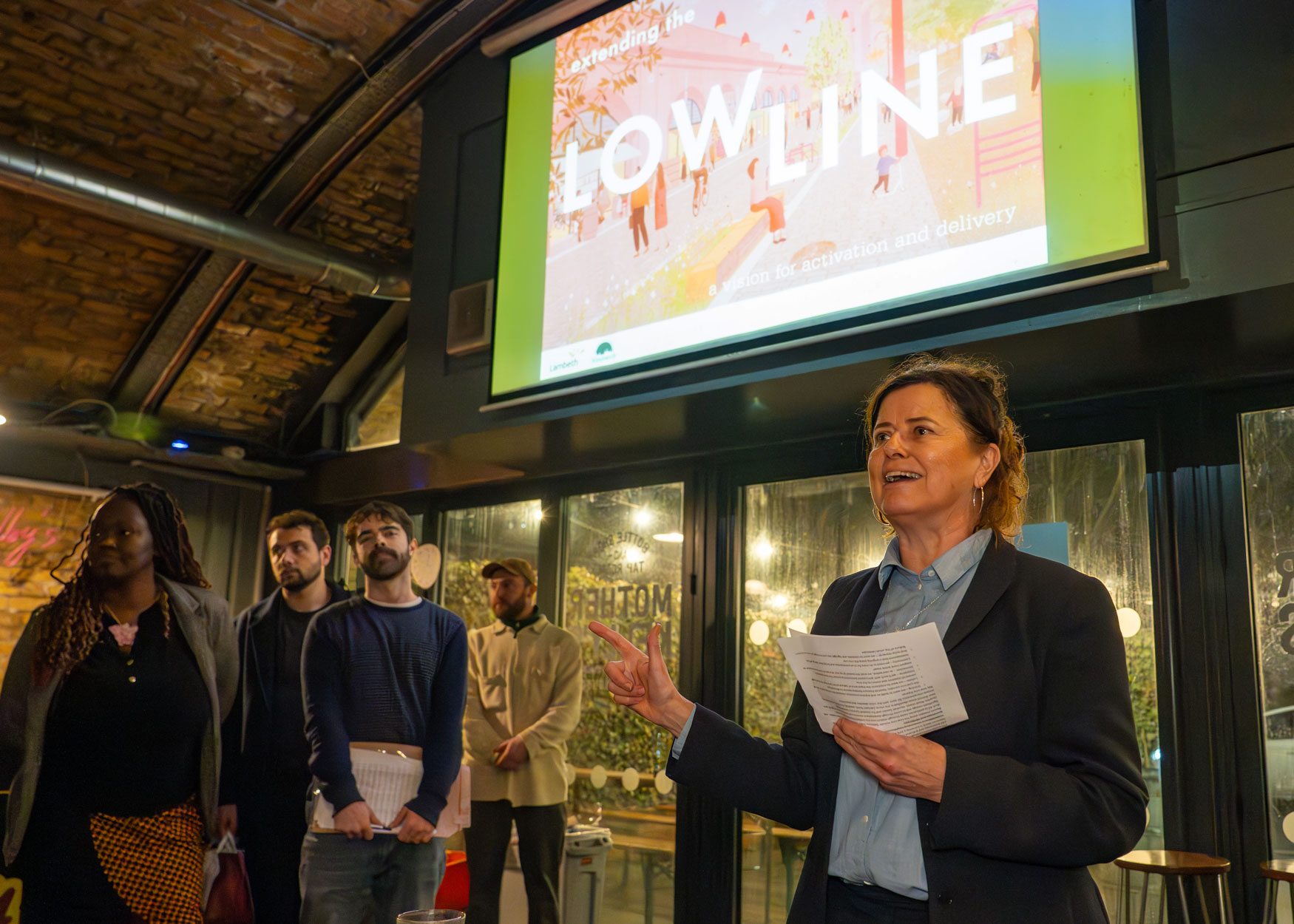 Lambeth's Leader, Councillor Claire Holland standing under a screen showing the cover of the Low Line Vision and speaking to a standing audience