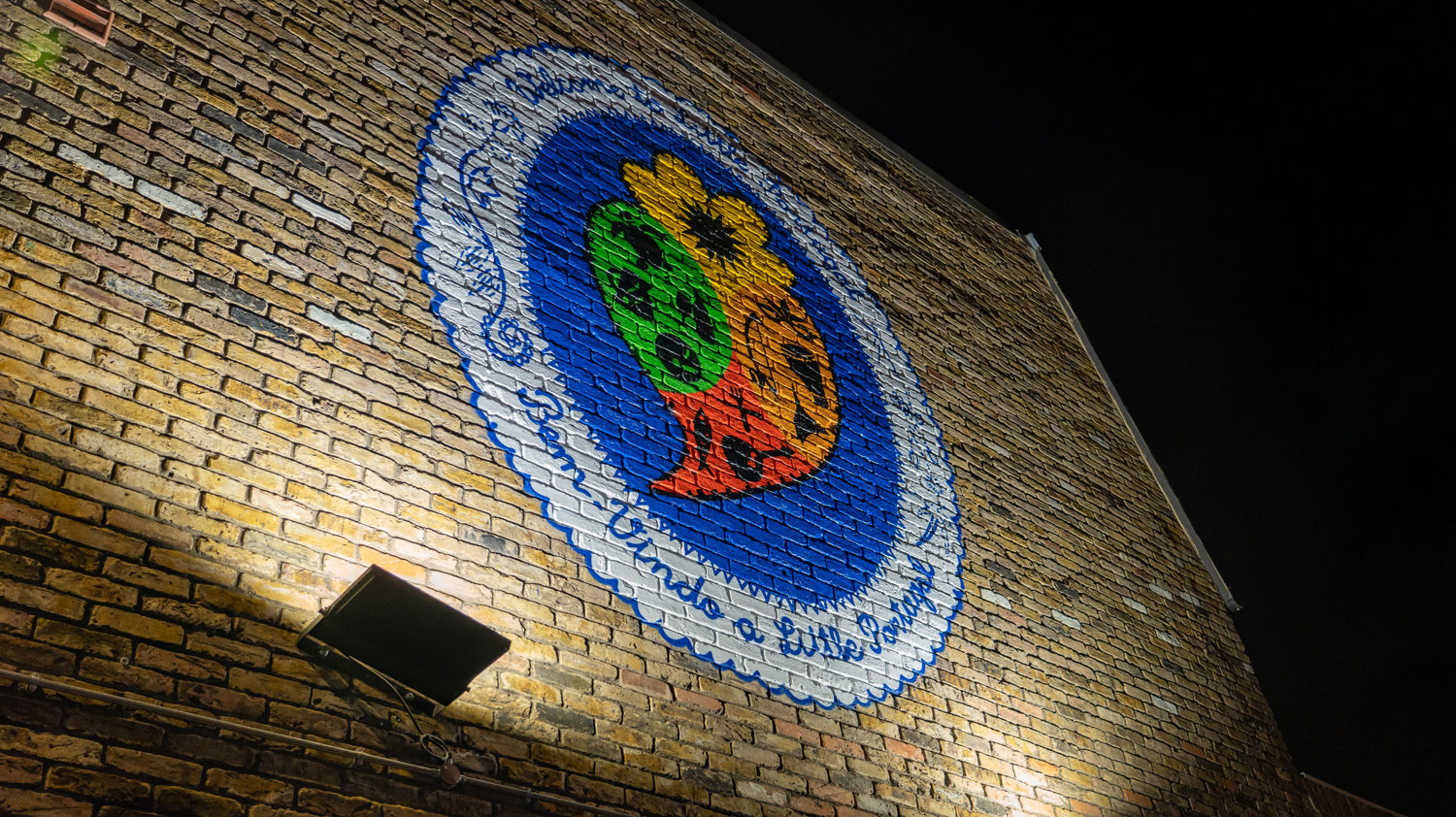 A photo of a mural painted on a brick wall, the mural is an oval shape with a white border reading "Welcome to Little Portugal" in English and Portuguese, and a stylised heart in the middle with colours and symbols representing Portuguese-speaking communities across the world