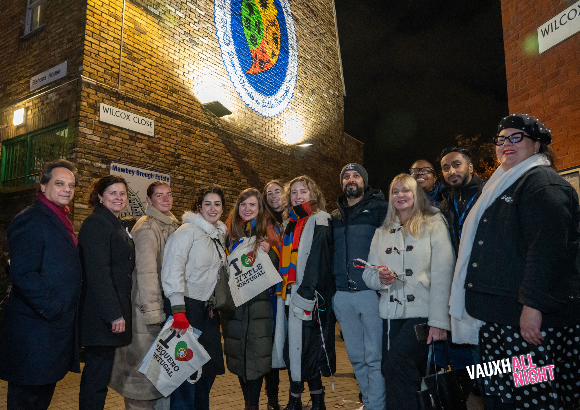 A group of people standing in front of a brick wall with a mural on it. The mural is an oval shape with a white border reading "Welcome to Little Portugal" in English and Portuguese, and a stylised heart in the middle with colours and symbols representing Portuguese-speaking communities across the world