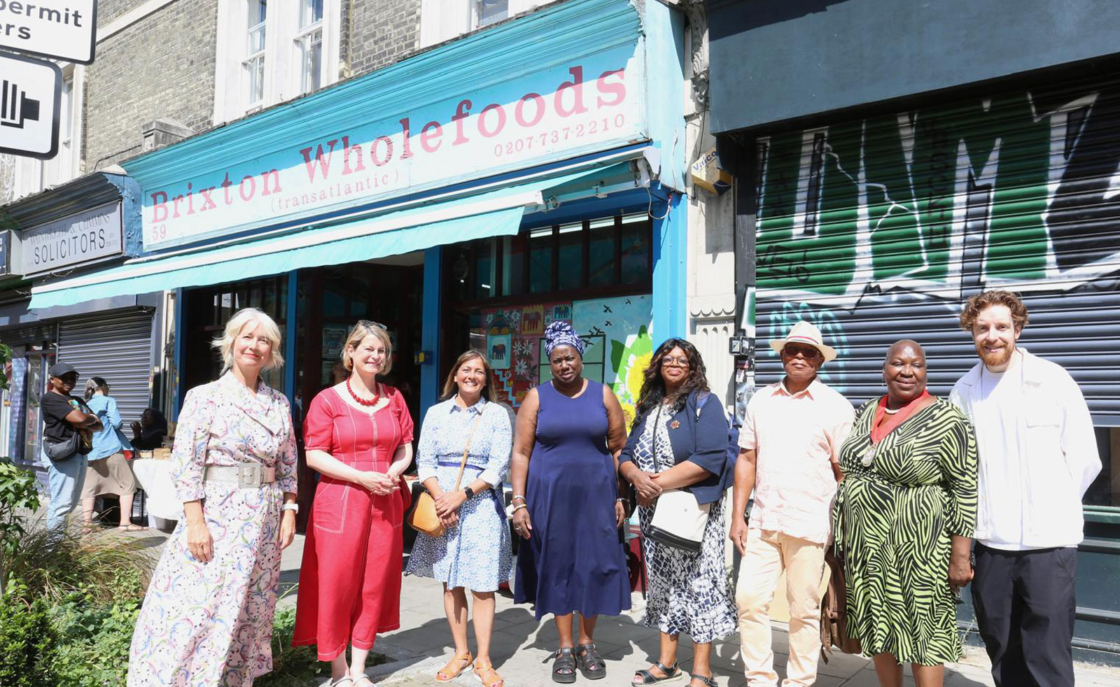 A group of Lambeth's Cabinet Members with local community members smiling in the sun outside Brixton Wholefoods on Atlantic Road