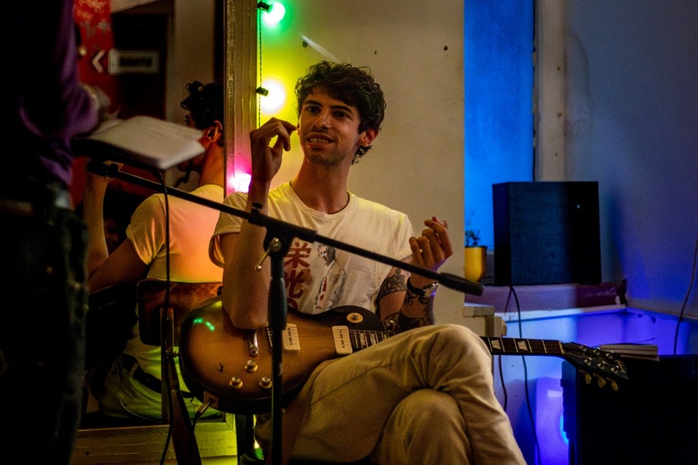 Male performer with brown hair, sat down holding a guitar at the KataKata community night time event