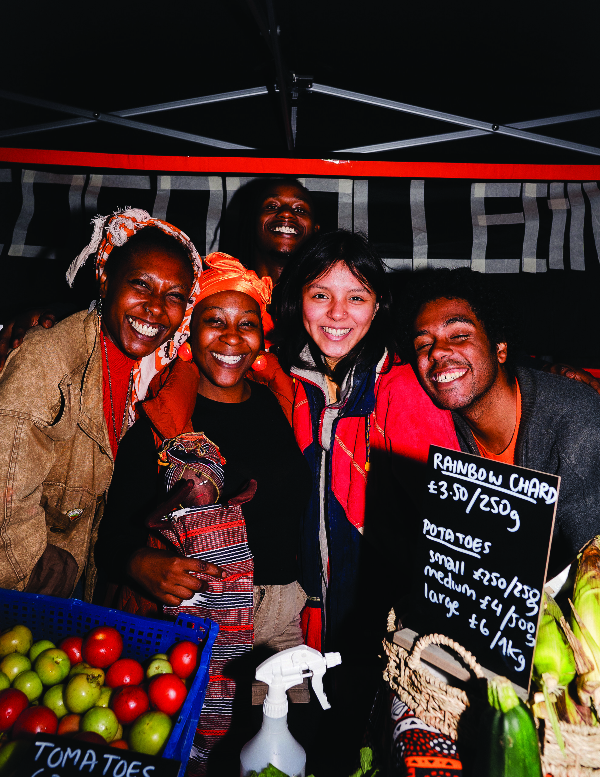 A group of five people standing under a gazebo and behind a table of food for sale, smiling at the camera