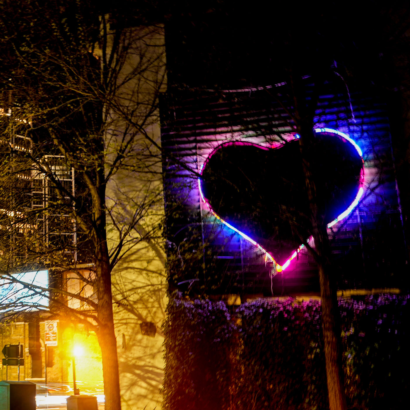 A night-time city scene in Vauxhall with illuminated buildings and a large neon heart artwork glowing on a wall, with trees and street lights in the foreground.