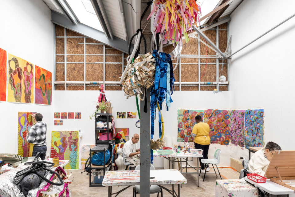 Artists work in a bright studio space, creating colourful paintings on canvases displayed on the walls, with materials and artwork spread across tables