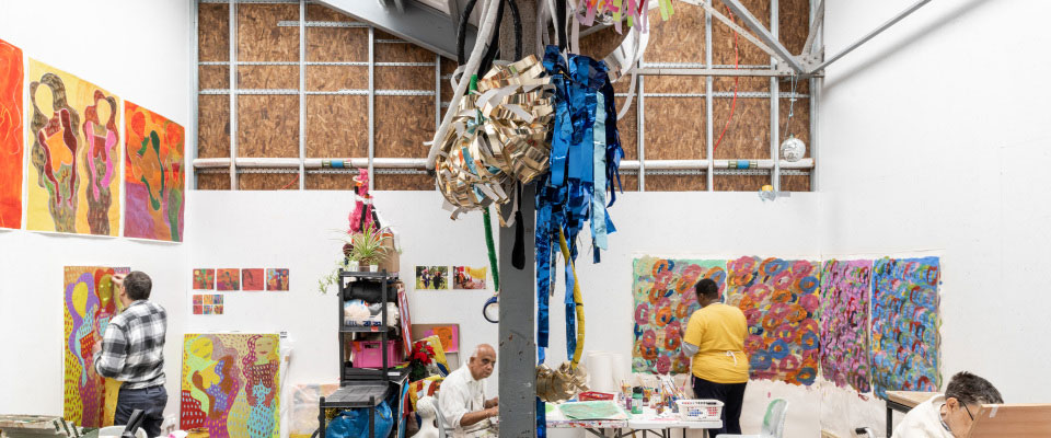 Artists work in a bright studio space, creating colourful paintings on canvases displayed on the walls, with materials and artwork spread across tables