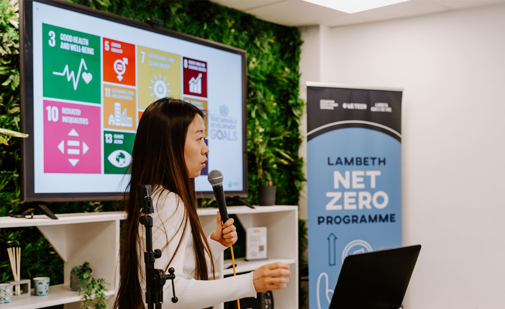 A person holding a microphone and presenting with a banner in the background which reads 'Lambeth Net Zero Programme'