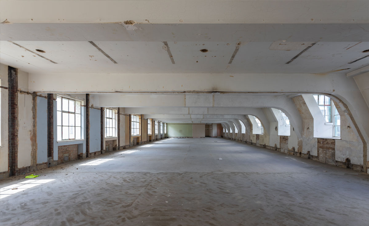 A large vacant space within a building with only a celling, windows and concrete floor.