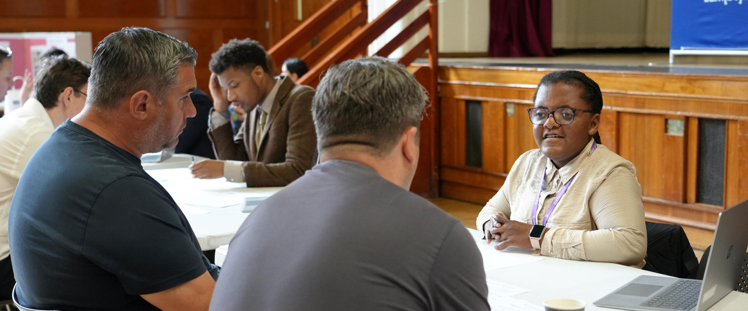 A woman speaks to two men across a table during a one-to-one meeting, with other participants seated and working in the background of a hall.