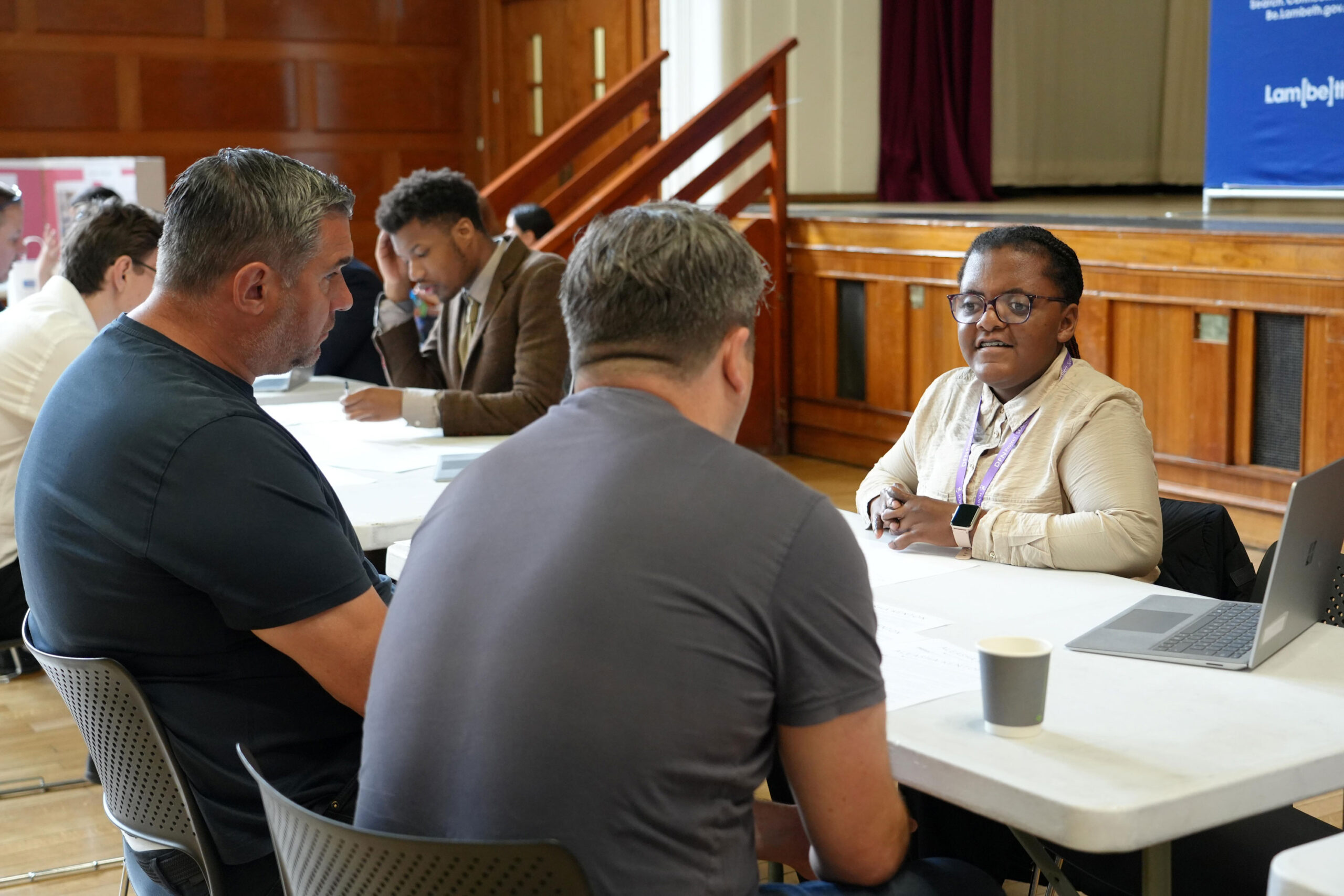 A woman speaks to two men across a table during a one-to-one advice session, with other participants seated and working in the background of a hall.