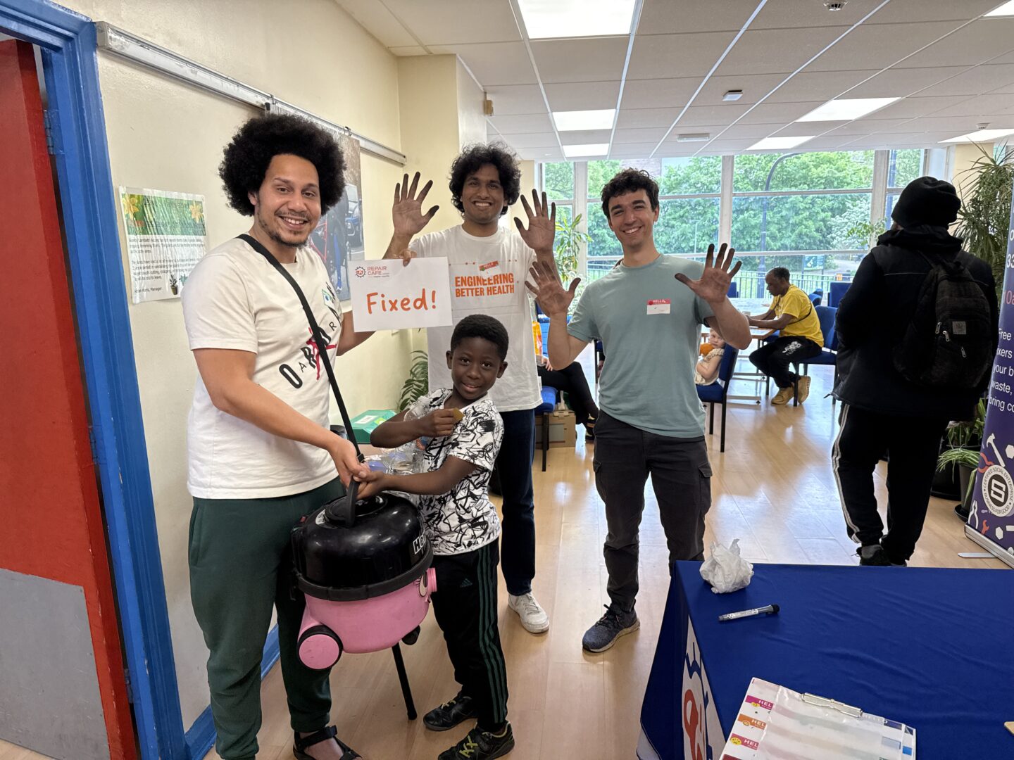 Members of the public and volunteers of the Repair Cafe pose with a vacum cleaner fixed during the session and hold a sign which reads 'Fixed!'