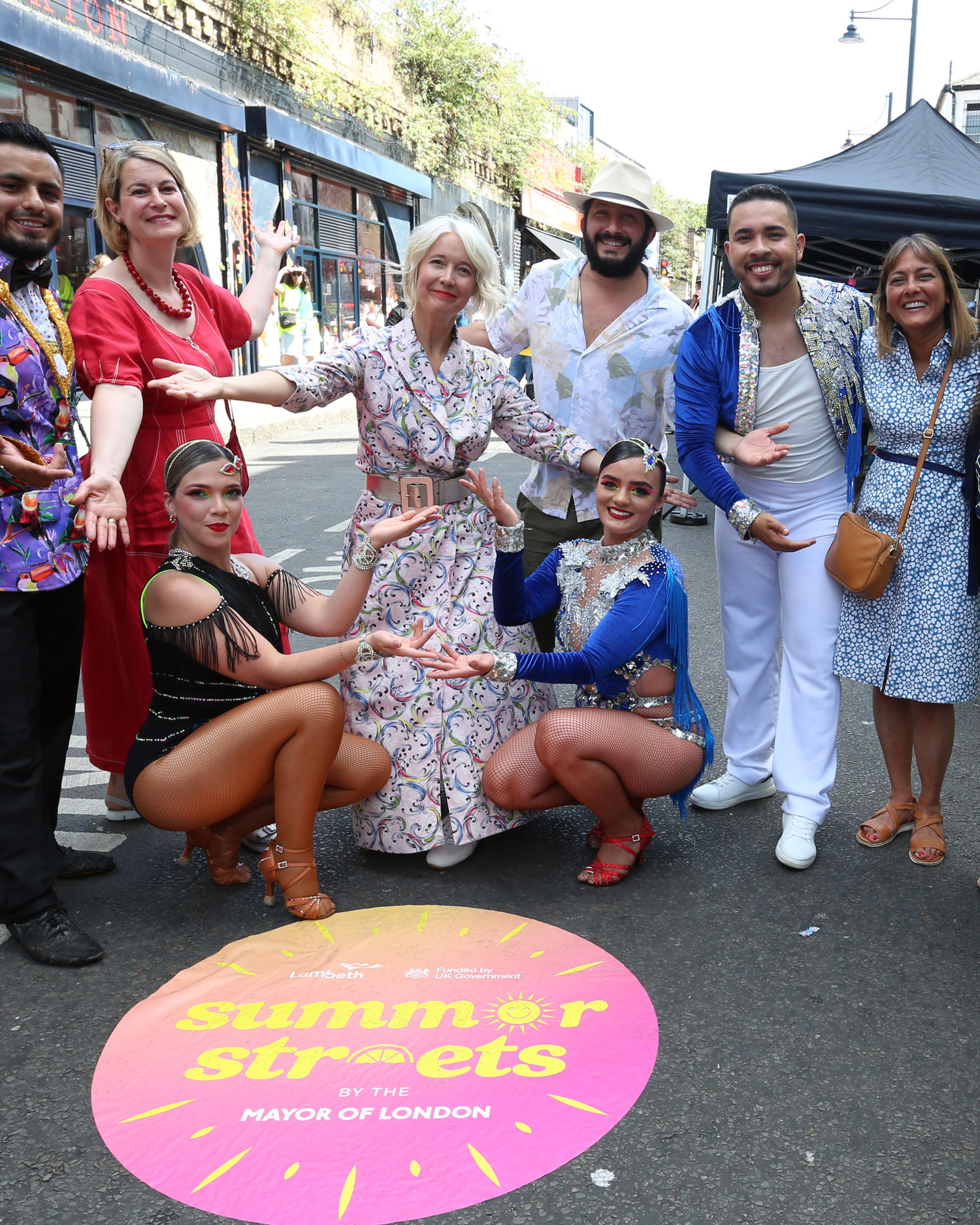 A group of people and performers in colourful outfits pose and smile on a closed street in Brixton during a Summer Streets event, with a sign on the road reading “Summer Streets by the Mayor of London.”
