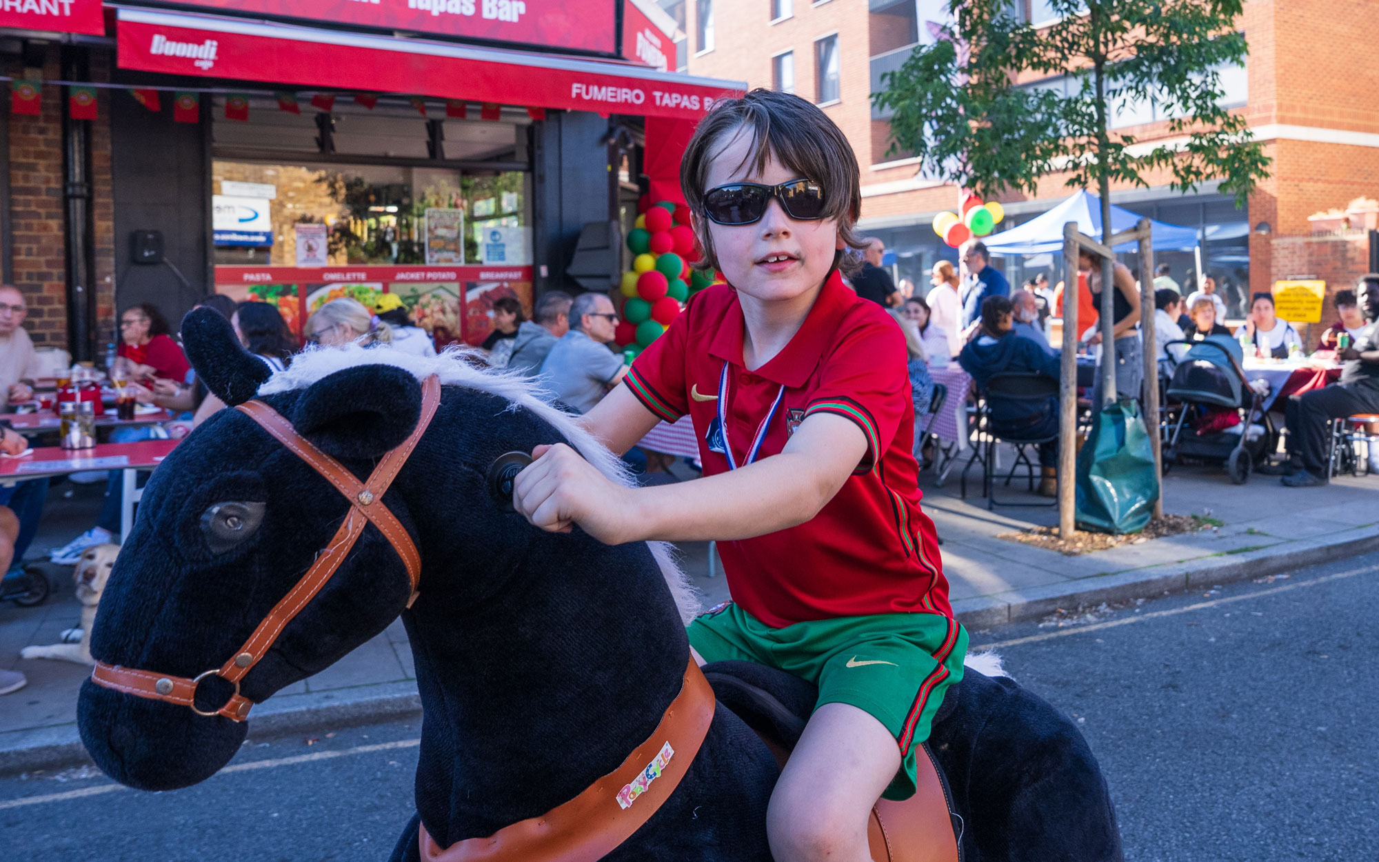 Child wearing a Portugal football kit rides a hobby horse through the festival street.