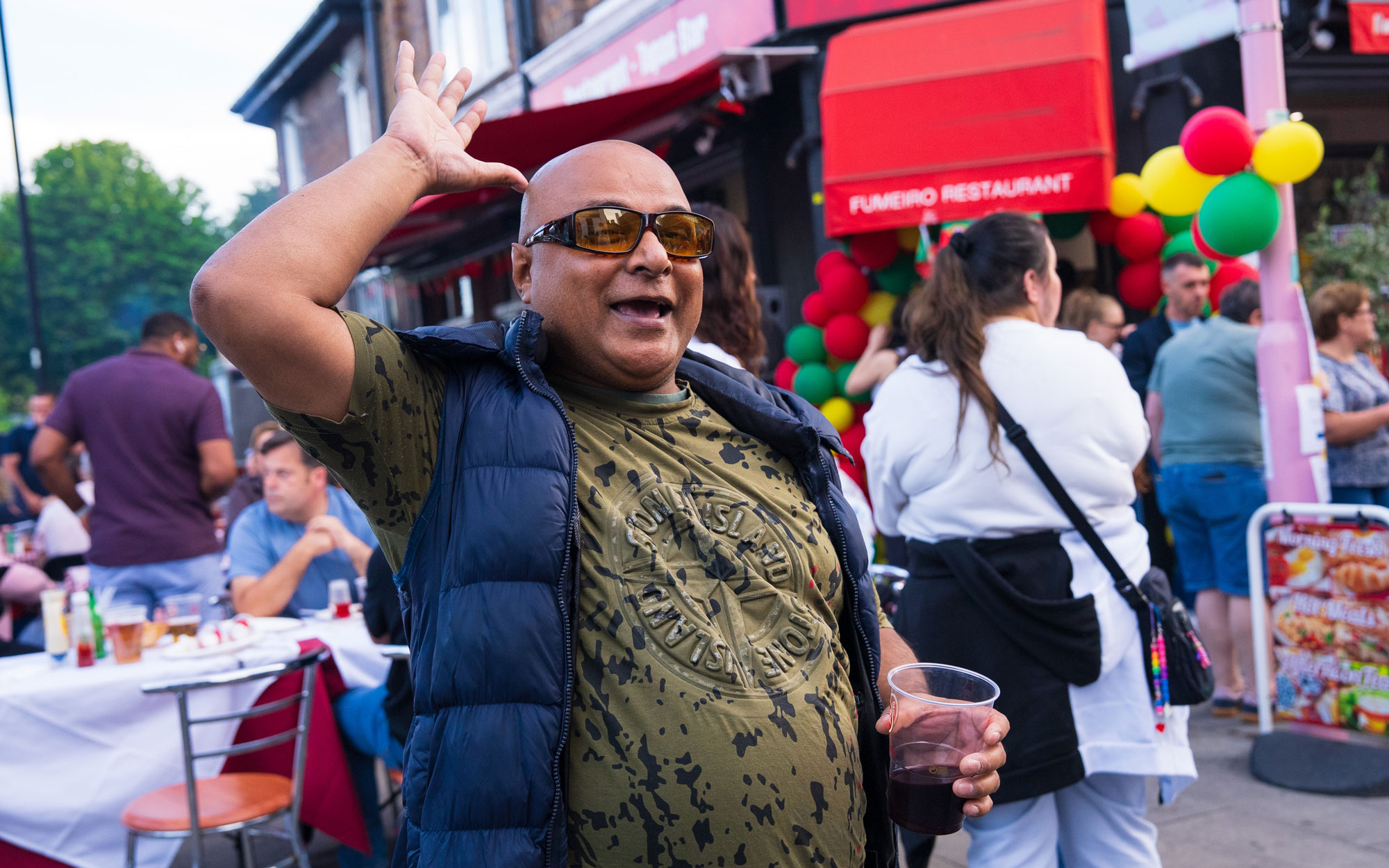 Festival-goer smiling and waving while holding a drink outside a Portuguese restaurant decorated for the event.