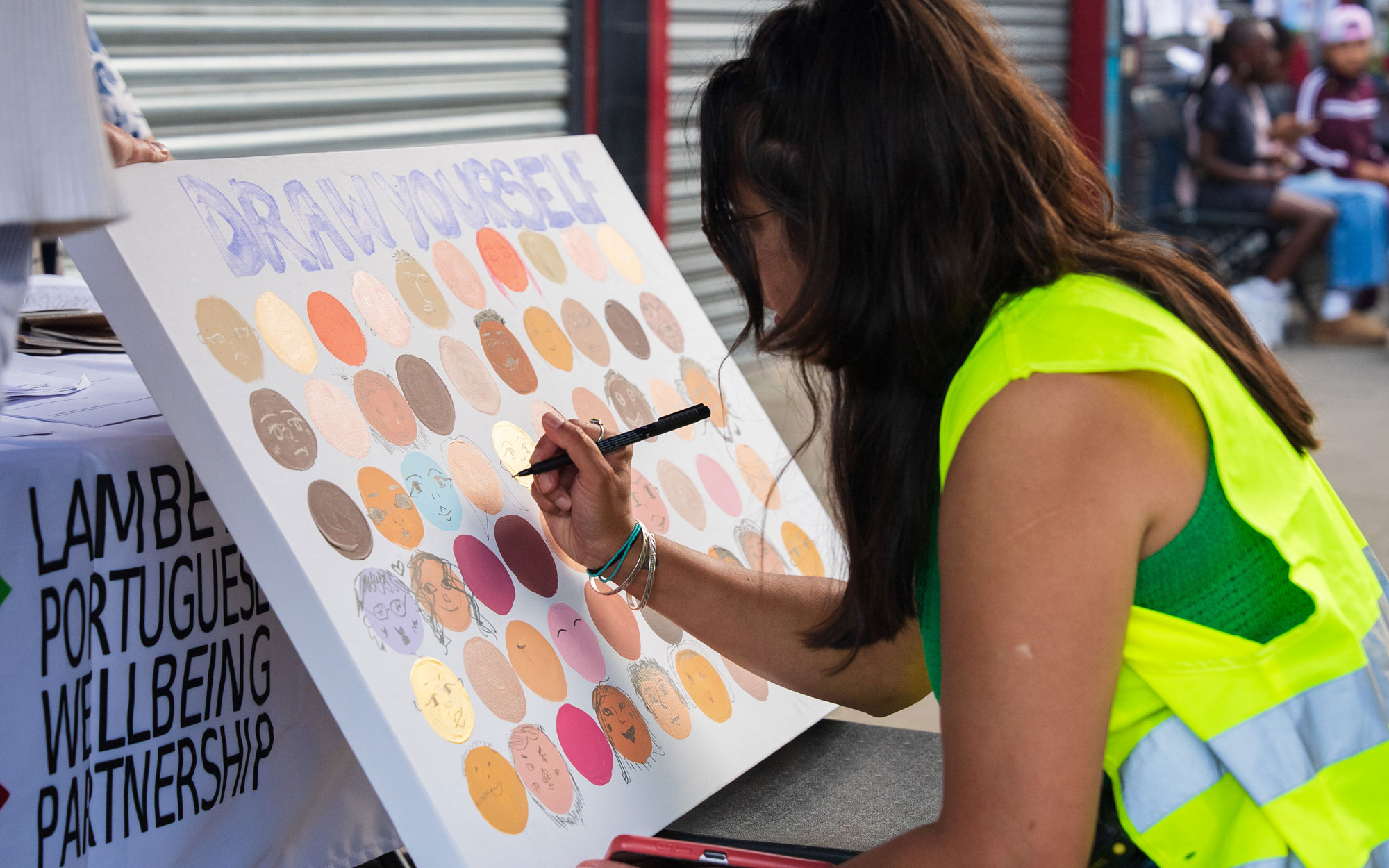 Participant adding a self-portrait to a collaborative community artwork at the Lambeth Portuguese Wellbeing Partnership stall.