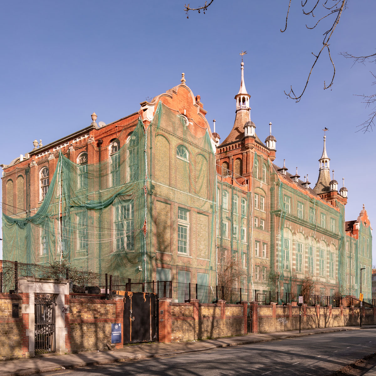 Street-level view of the historic former Cormont Road School building wrapped in green protective netting during restoration works on a clear sunny day.
