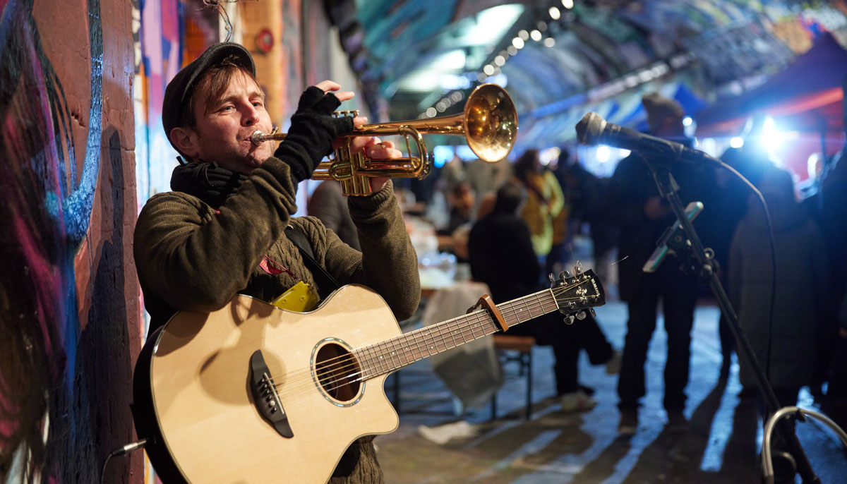 A street musician plays trumpet and guitar in an illuminated tunnel at night, with people gathered and walking in the background.