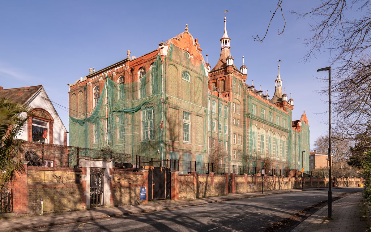 Street-level view of the historic former Cormont Road School building wrapped in green protective netting during restoration works on a clear sunny day.