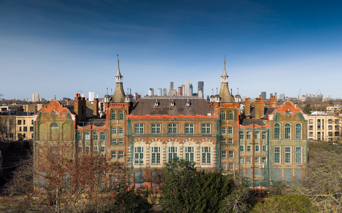Front view of the historic former Cormont Road School building with decorative towers and brickwork, partially covered in green protective netting, with central London skyline visible behind.