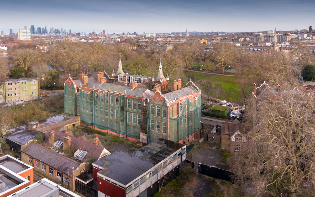 Aerial view of the historic former Cormont Road School building in Lambeth, partially covered in green protective netting, surrounded by neighbouring homes, trees and the London skyline in the distance.