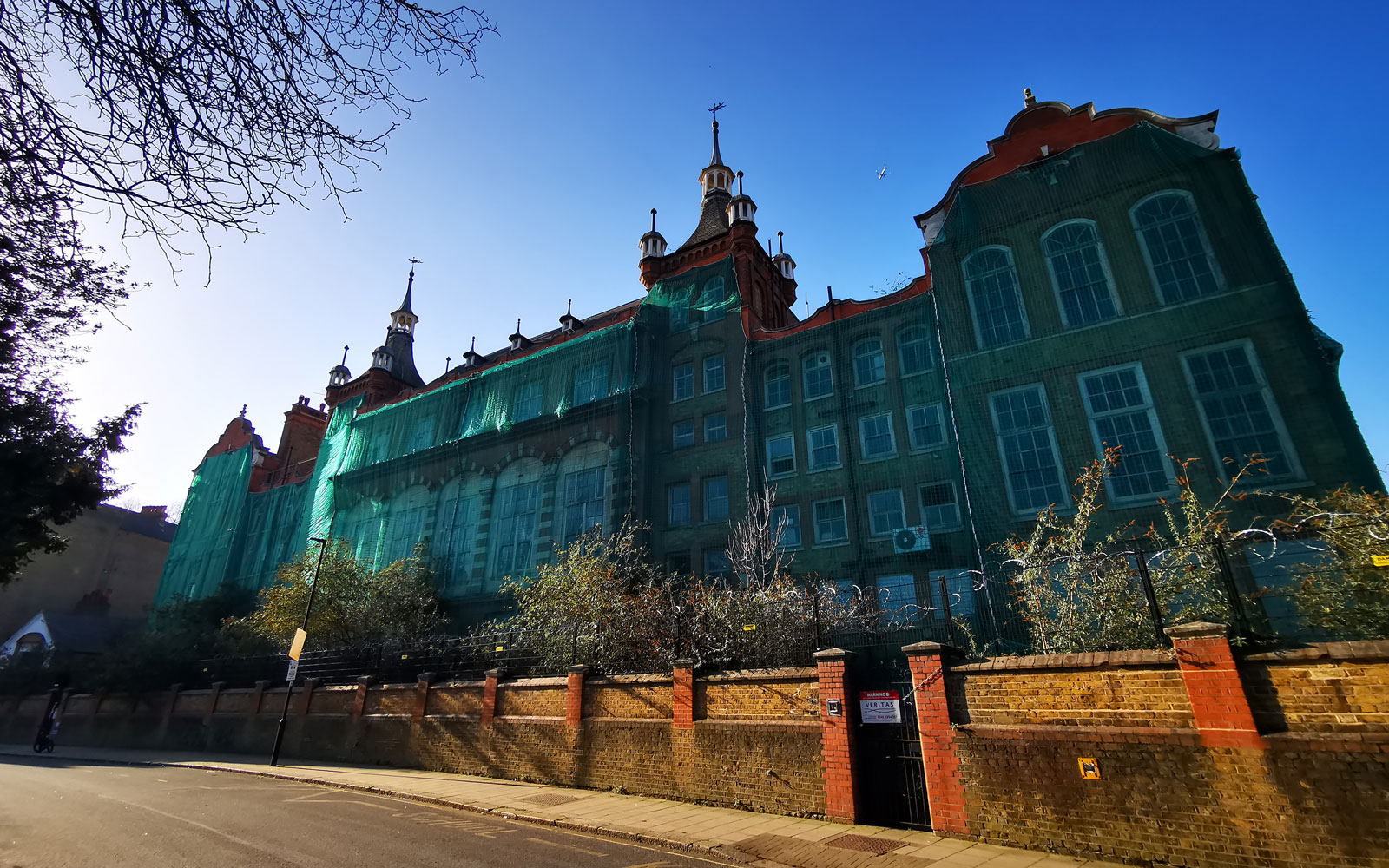 Historic former school building covered in green protective netting during restoration works, viewed from the street on a clear sunny day.