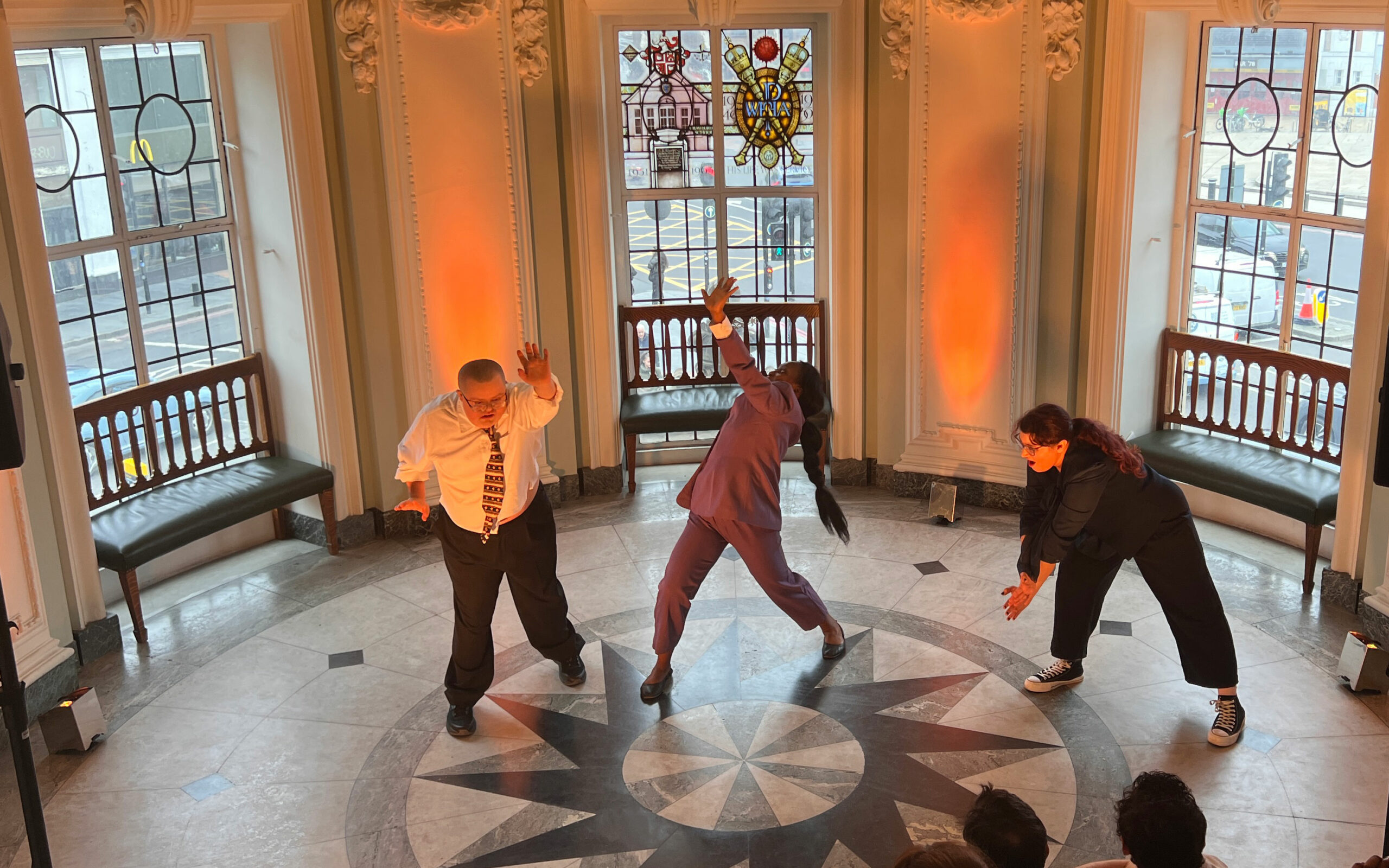 Three performers dance together in the centre of Lambeth Town Hall's entrance hall during a Danceology performance, with the audience watching from above.