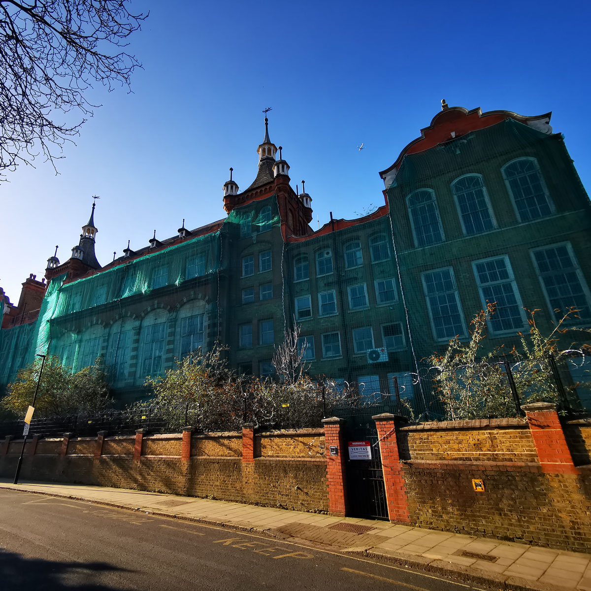 Historic former school building covered in green protective netting during restoration works, viewed from the street on a clear sunny day.