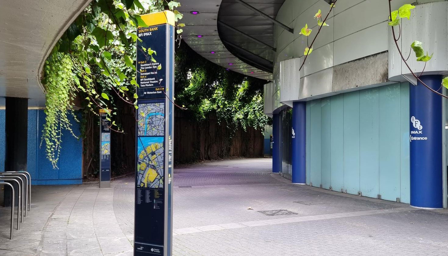 Image 1 alt text: A pedestrian walkway near the BFI IMAX on the South Bank, with wayfinding signs, greenery hanging overhead and curved modern buildings lining the route.