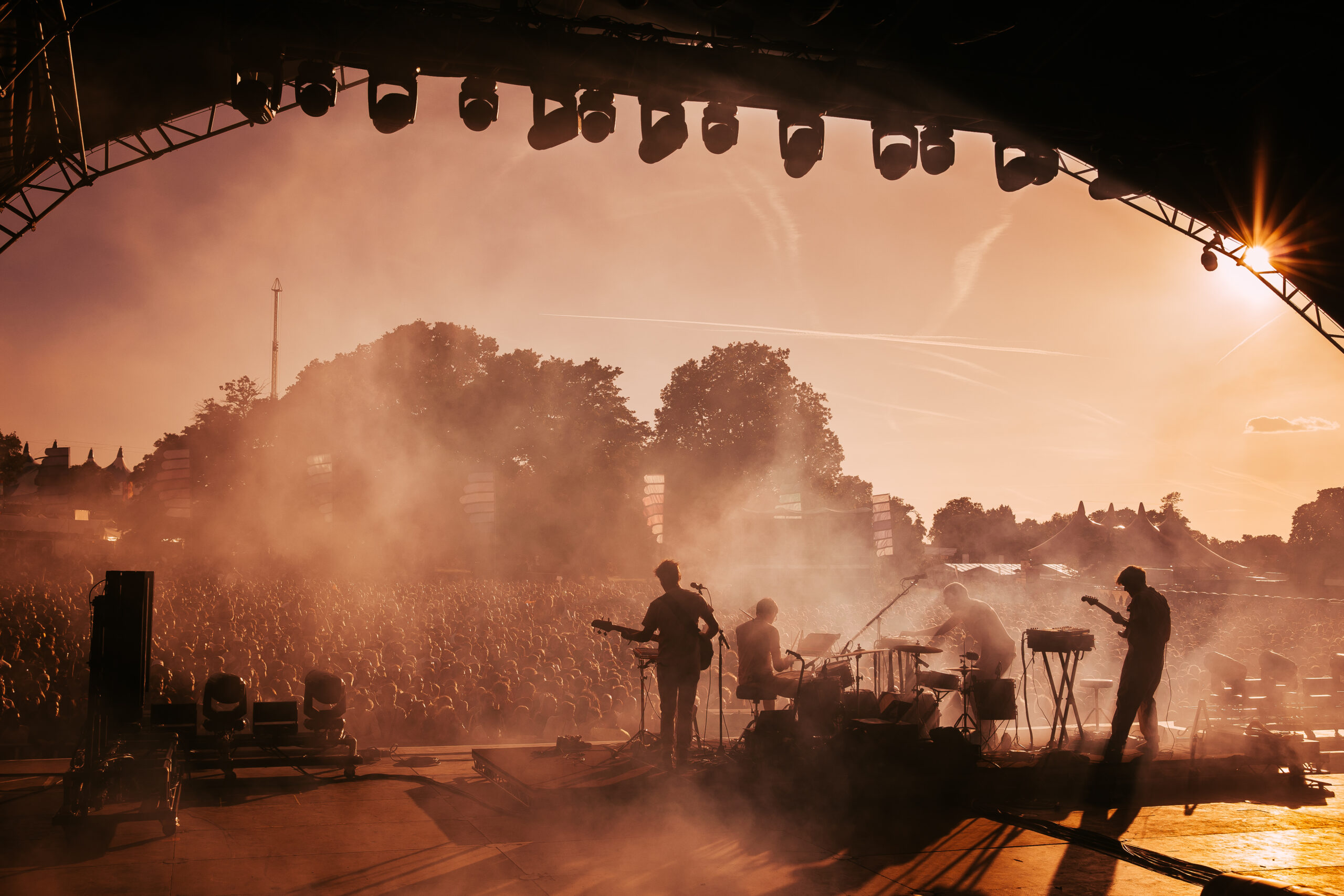 Musicians perform on stage to a large crowd at an outdoor music festival at sunset.