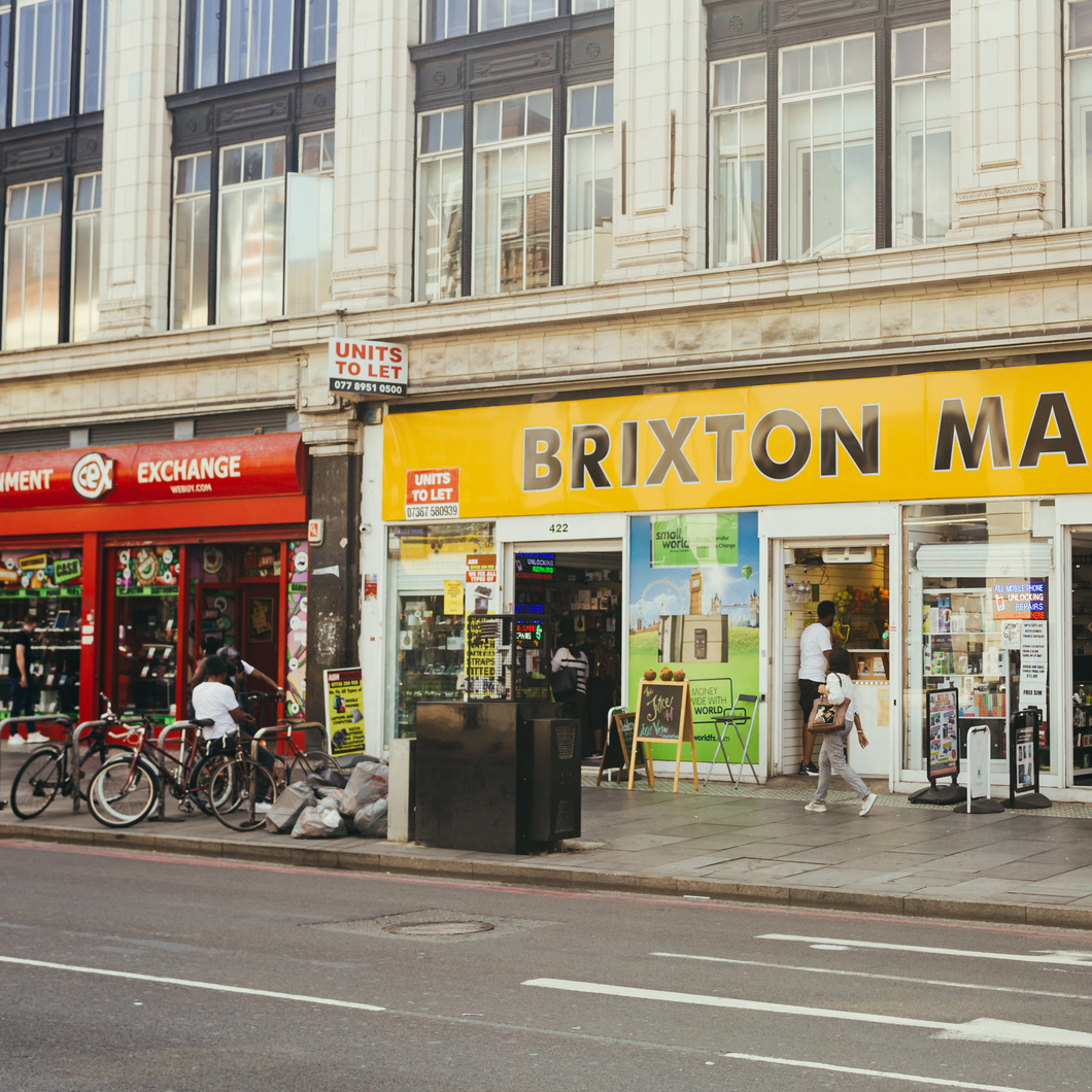 Street-level view of Brixton Mall on Brixton Road, showing independent shops, pedestrians and cyclists outside the entrance to the busy indoor market.