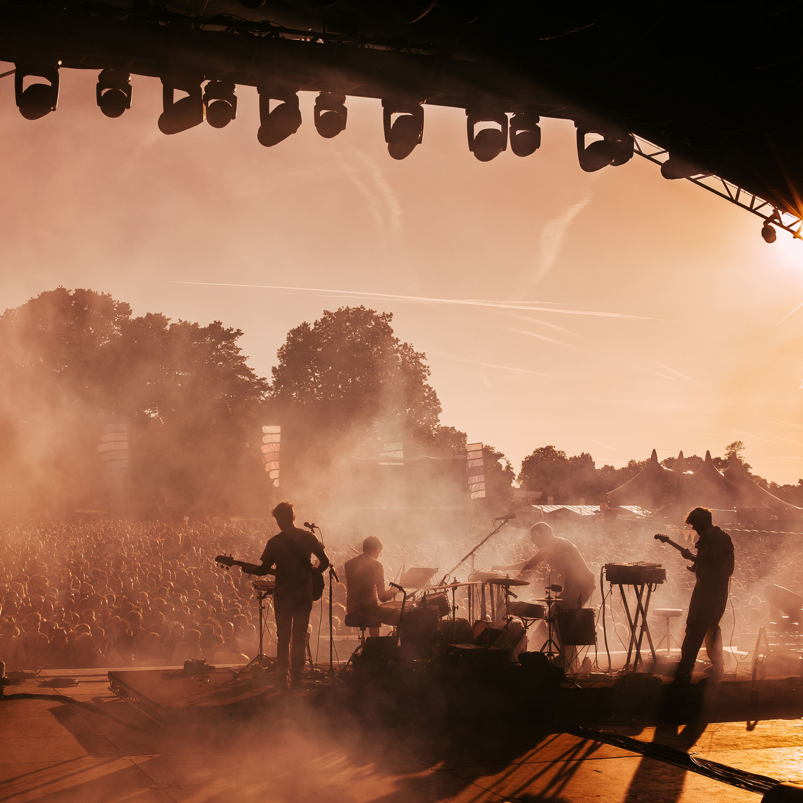 Musicians perform on stage to a large crowd at an outdoor music festival at sunset.