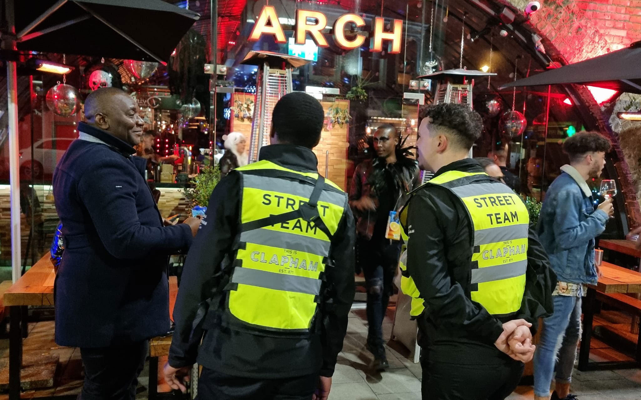 Clapham street team members in high-visibility jackets speak with visitors outside a busy venue in Clapham at night.