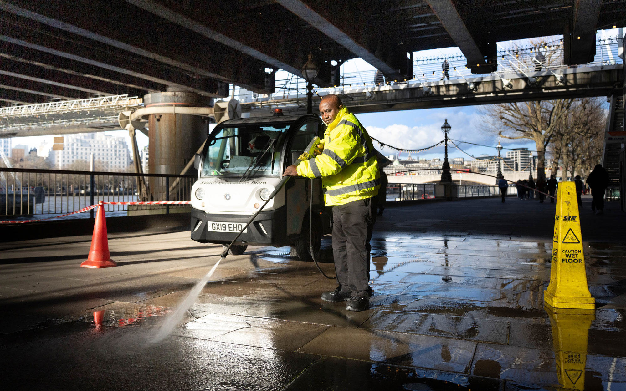South Bank cleaning team member pressure-washes the riverside walkway beneath a bridge using a maintenance vehicle and jet washer.