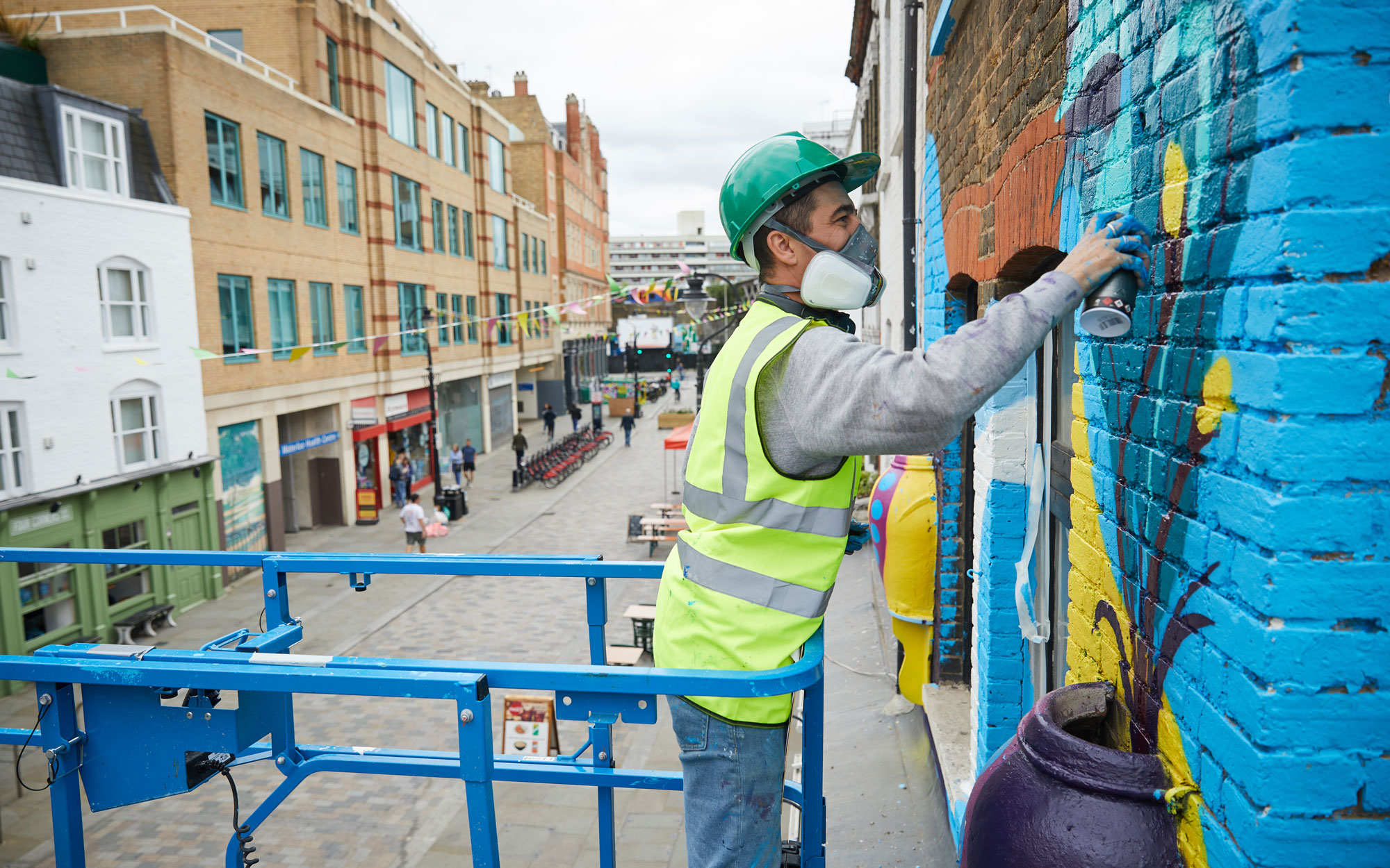 Artist wearing safety equipment spray-paints a colourful mural from a raised platform overlooking a pedestrian street in Waterloo.
