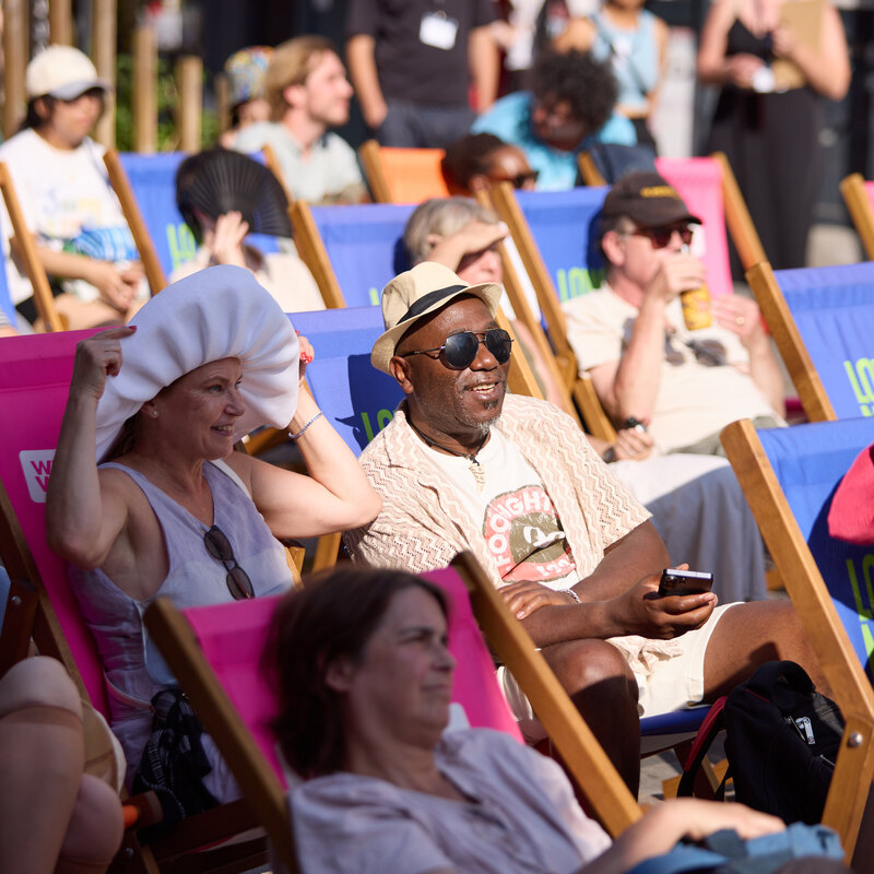 A group of people wearing summer clothing sit in pink and blue deckchairs outdoors on the street