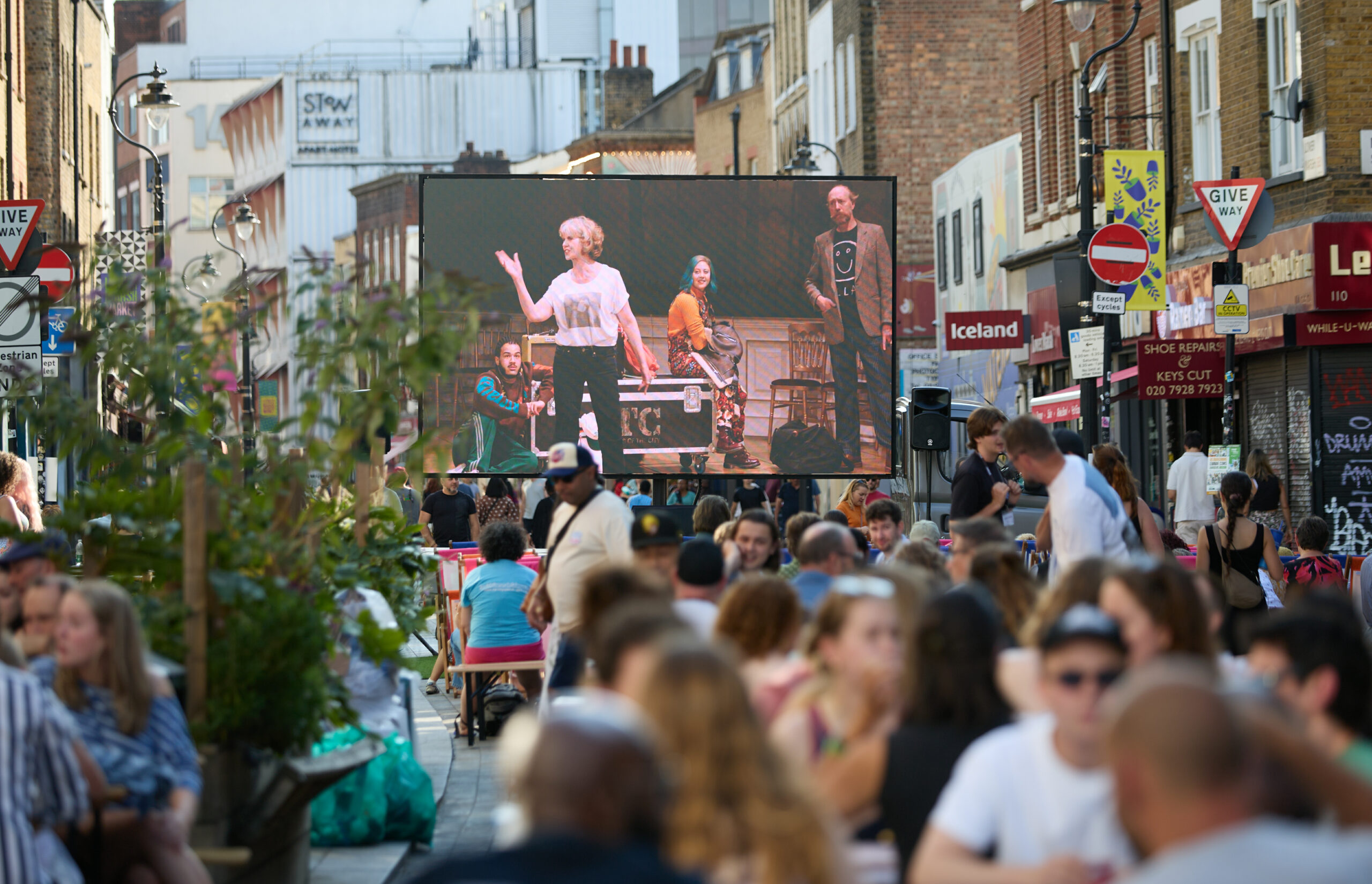 A photo of a large outdoor film screen set up on a busy street. People are sat in deckchairs watching the screen, while others sit at picnic tables.