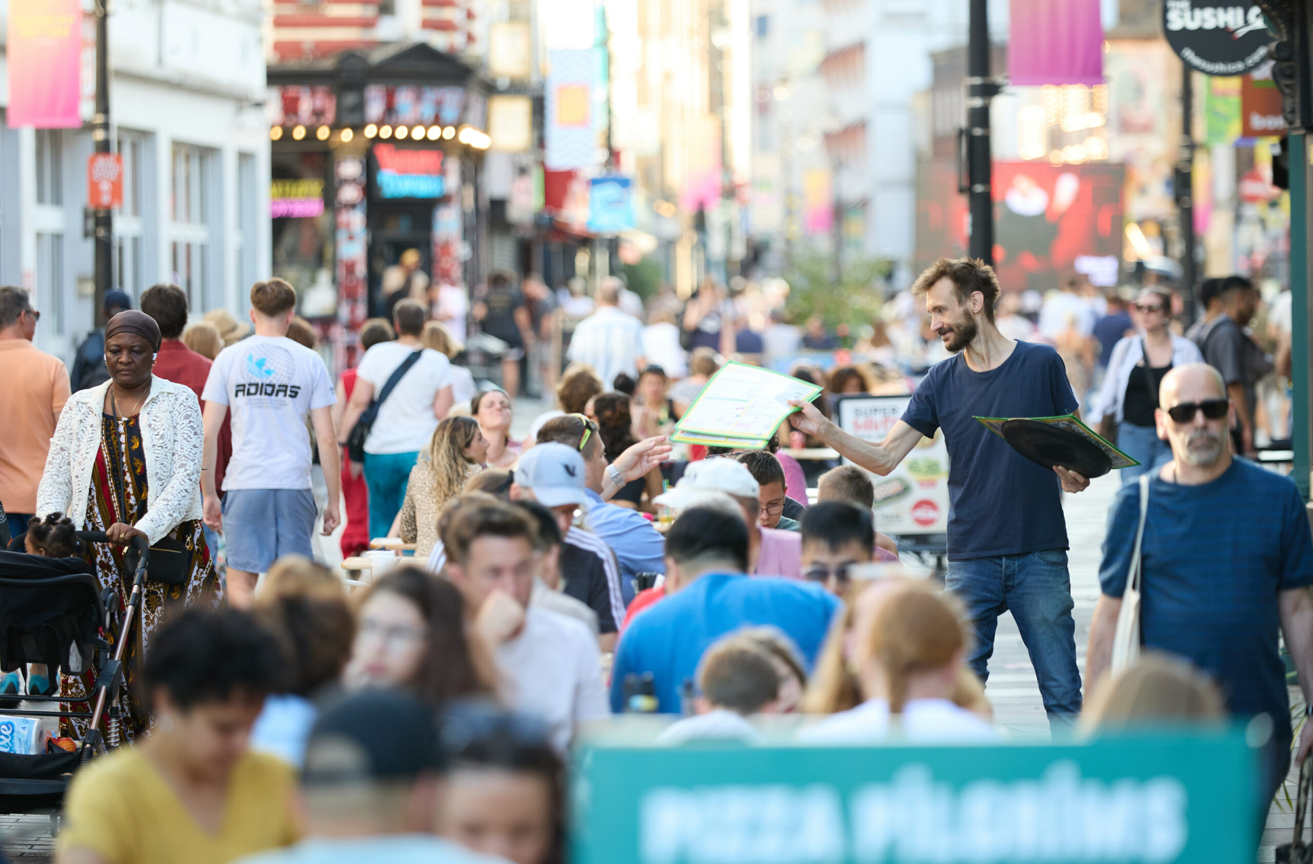 A waiter in a navy blue t-shirt hands out menus to a group of people sitting at outdoor tables on a busy street