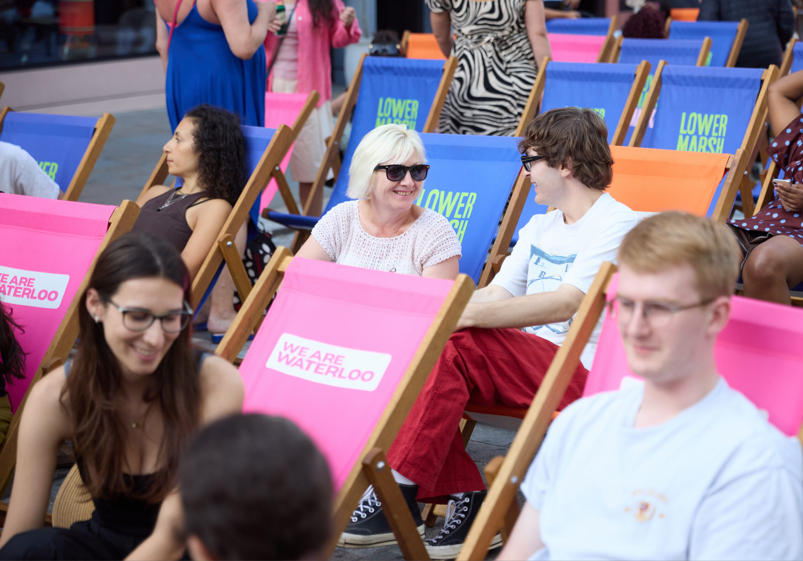 A group of people sit in pink, blue and orange deckchairs outdoors in a street, smiling and talking