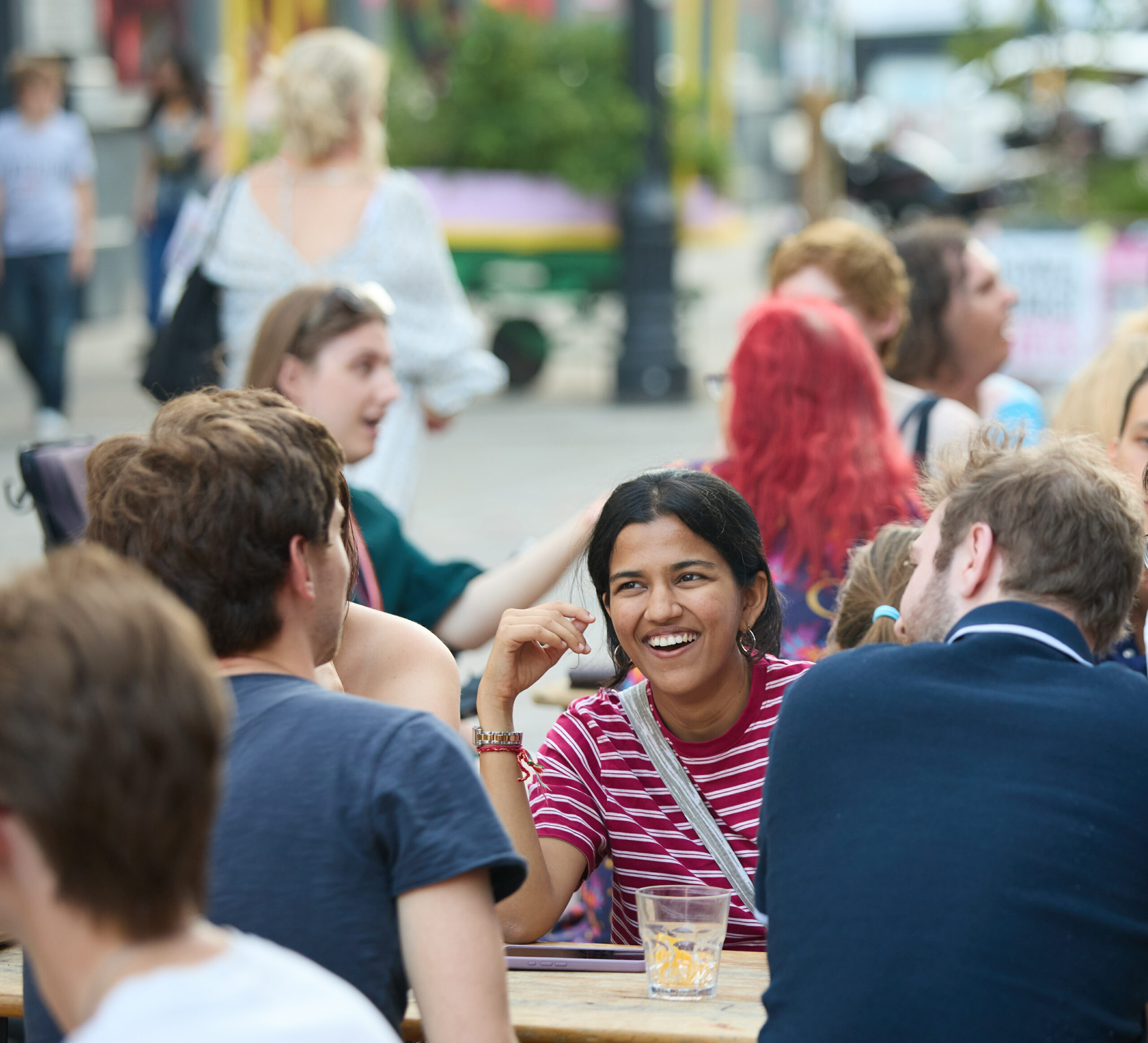 A person sitting at an outdoor table is smiling and talking to friends sat across from them. A beverage is on the table in front of them.