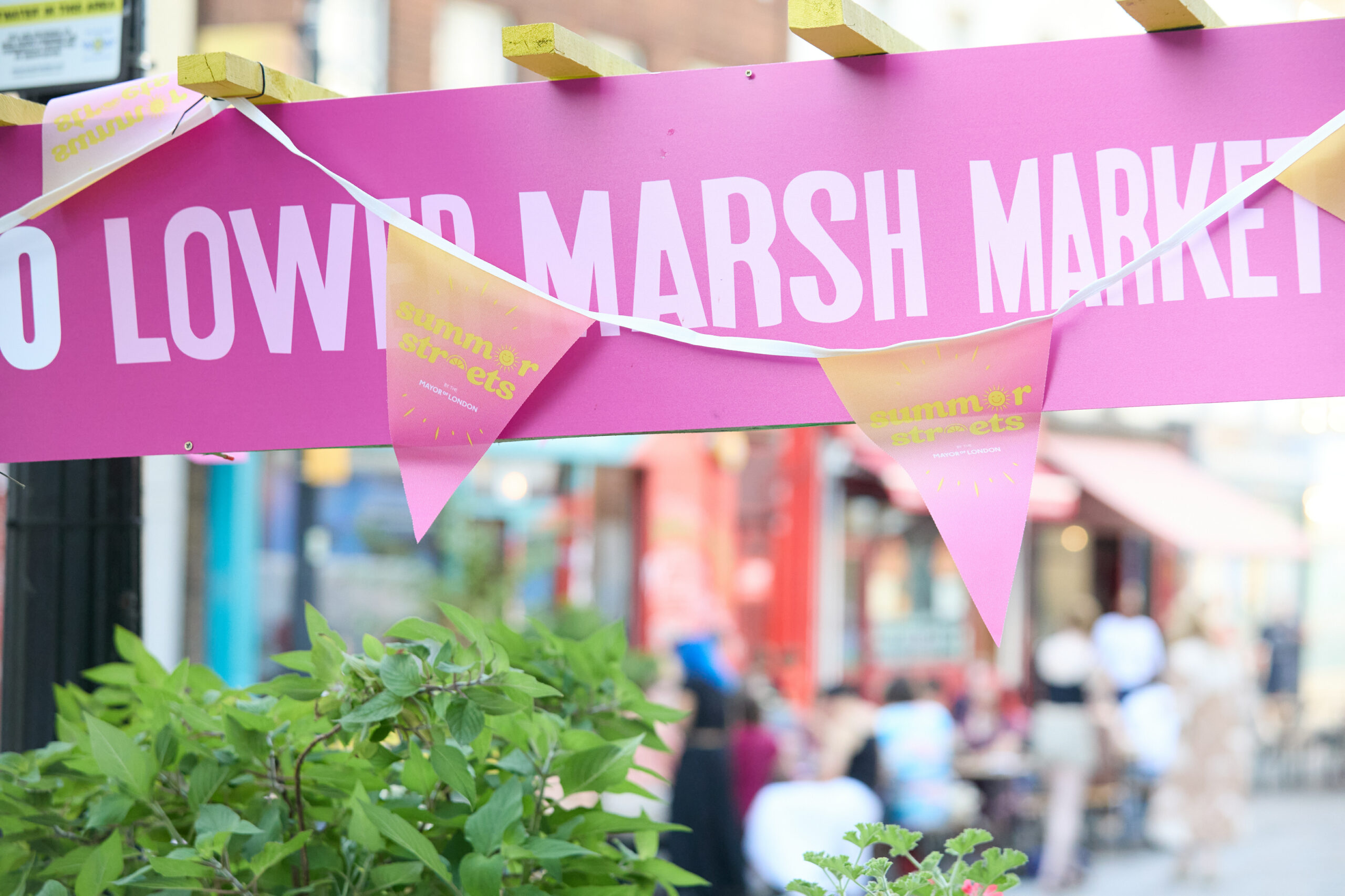 A photo of a pink awning reading "Lower Marsh Market" in white. Pink bunting reading "Summer Streets" in yellow is hanging over the awning. Green plants are at the bottom.