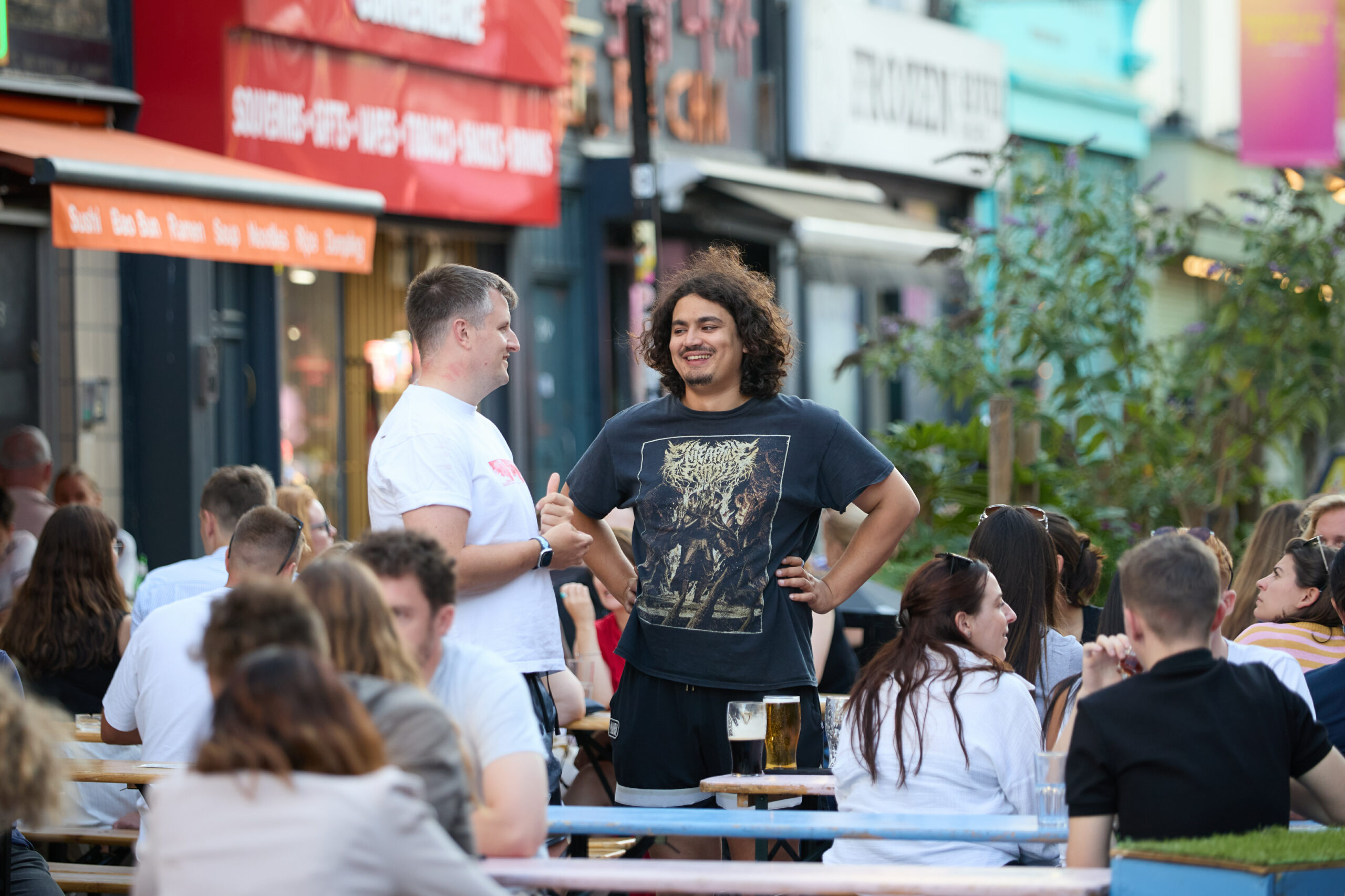 Two people standing up and chatting in the midst of picnic tables set up outdoors on a busy street