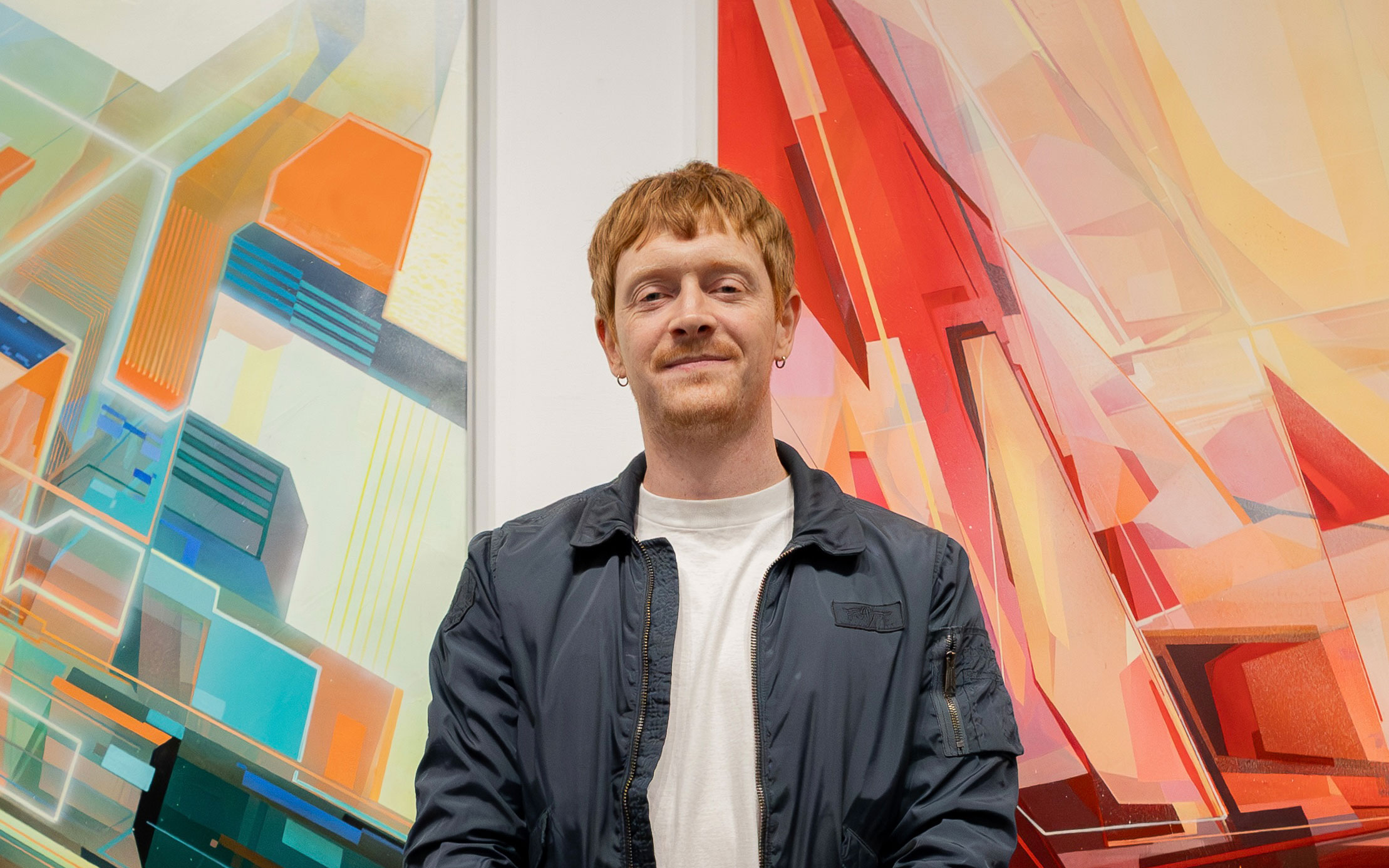 Artist Jake Attewell stands between two colourful abstract paintings at a gallery, wearing a navy jacket and a white T-shirt