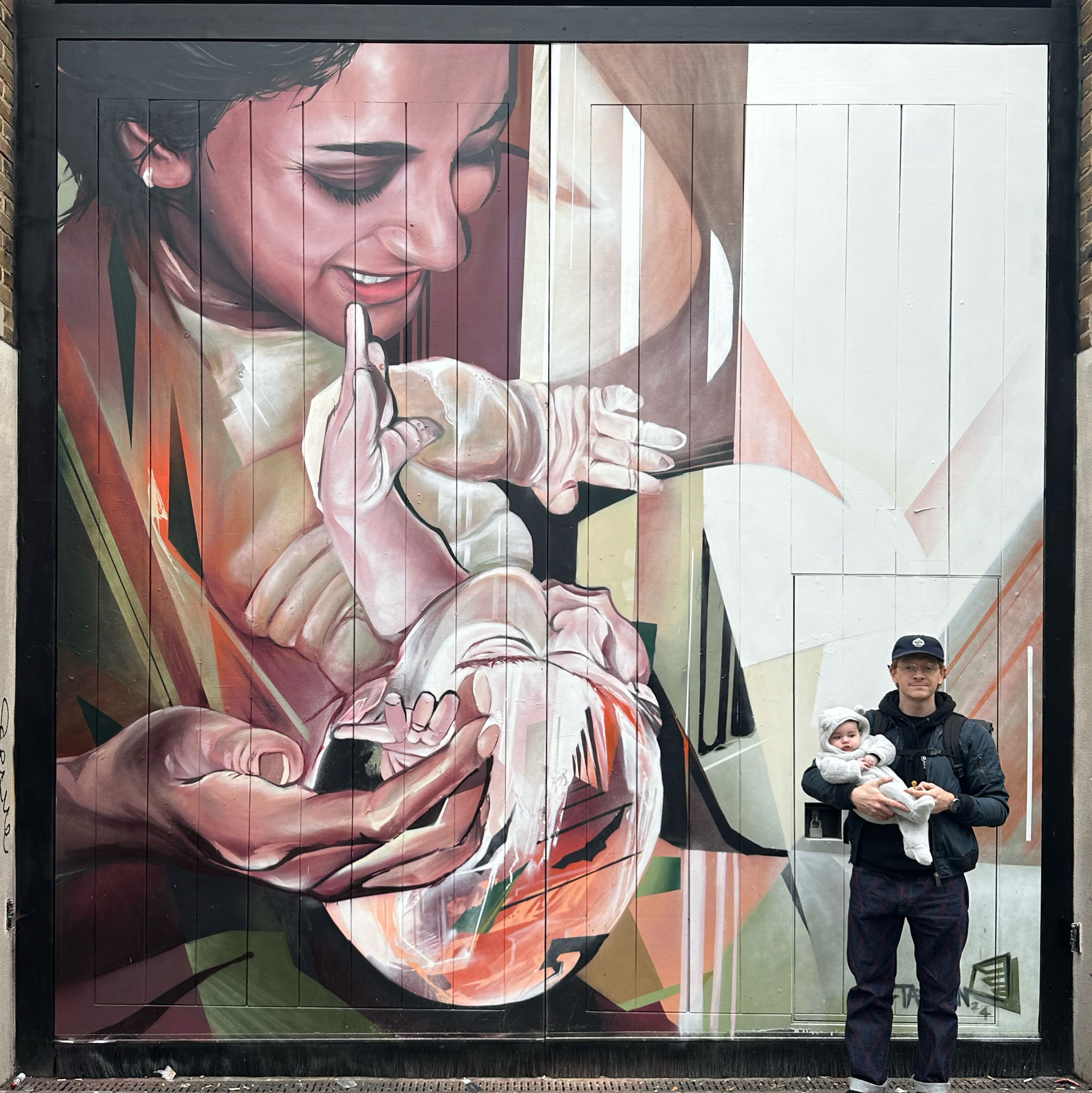 Artist Jake Attewell stands holding a baby in front of one of his large murals depicting a parent cradling a child in a blend of realism and abstract forms.